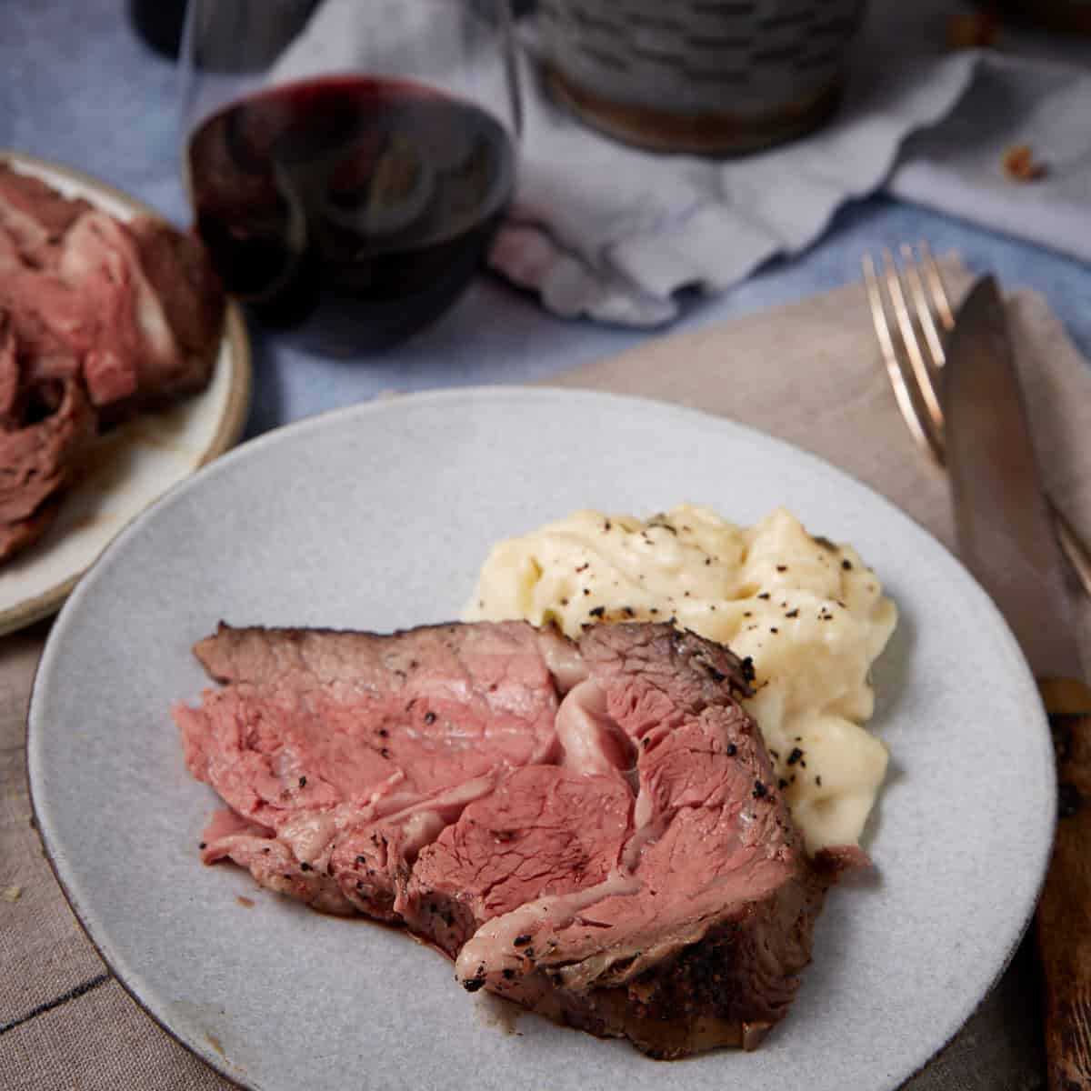 A plate with a thick slice of medium-rare standing rib roast and a serving of mashed potatoes, sprinkled with black pepper. In the background, there is a glass of red wine and cutlery on a napkin.