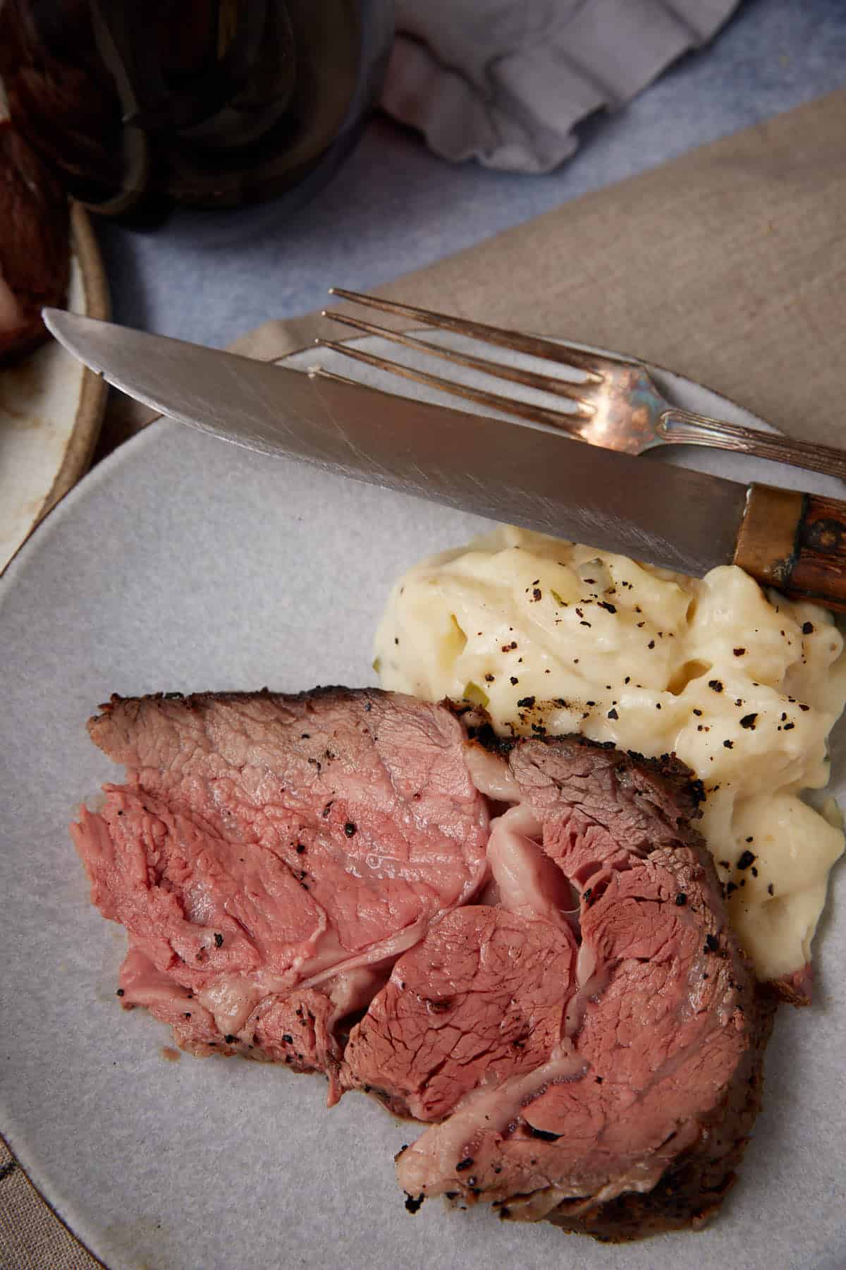 A plate with two slices of medium-rare standing rib roast and a serving of creamy mashed potatoes, topped with cracked black pepper. A knife and fork rest on the plate.