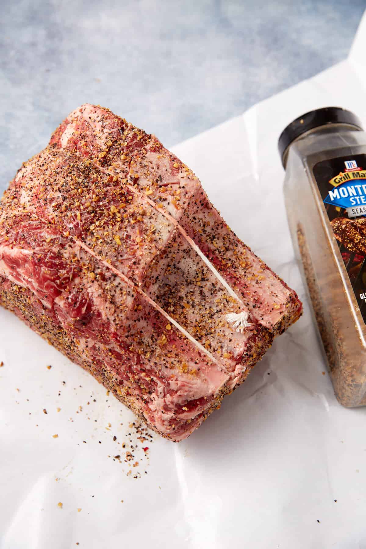 A raw, seasoned standing rib roast is tied with string and sits on white parchment paper next to an open container of Montreal steak seasoning.