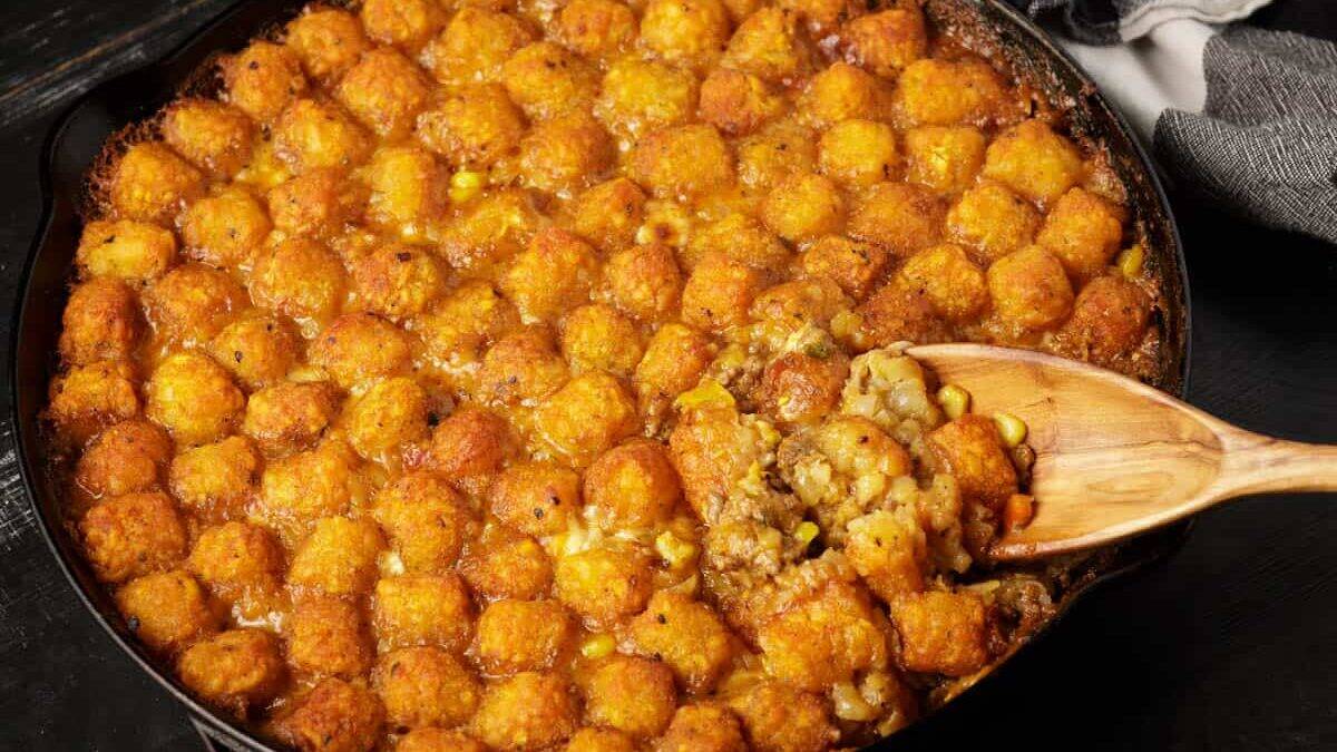 A skillet filled with golden-brown tater tot casserole, with a wooden spoon scooping out a portion to reveal a filling of vegetables and sauce underneath the crispy tater tot topping.