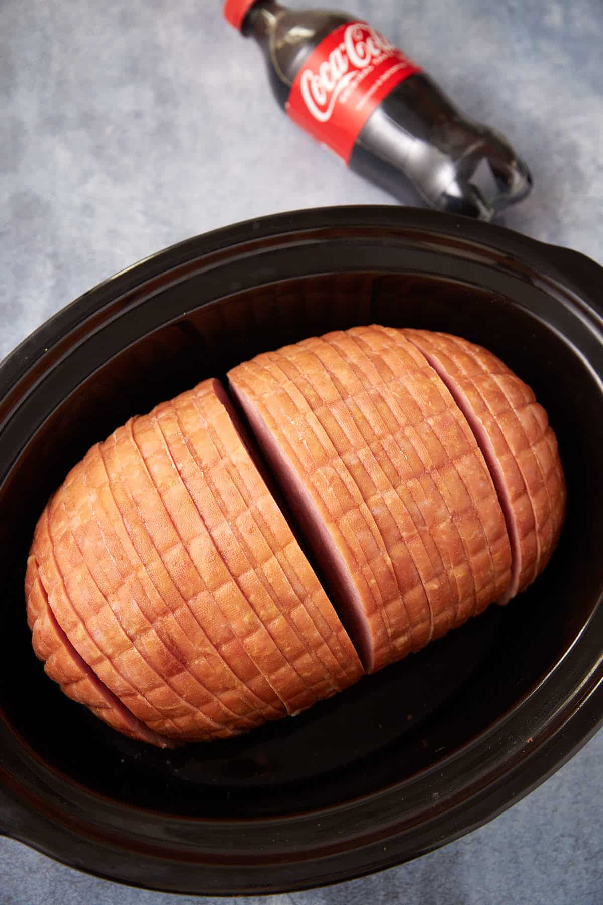 A whole, sliced ham placed in a black slow cooker, with a bottle of Coca-Cola lying on its side in the background.