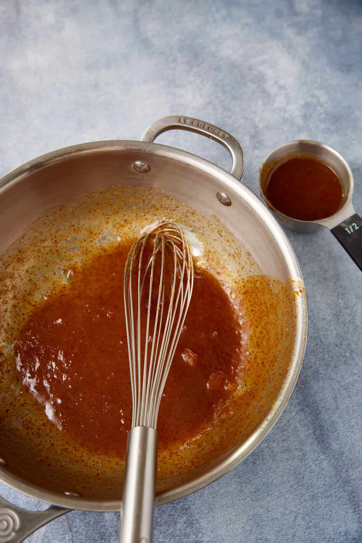 A metal saucepan containing a whisk and a thick reddish-brown sauce sits on a blue surface. Beside it, there is a metal half-cup measuring cup with more of the same sauce inside.