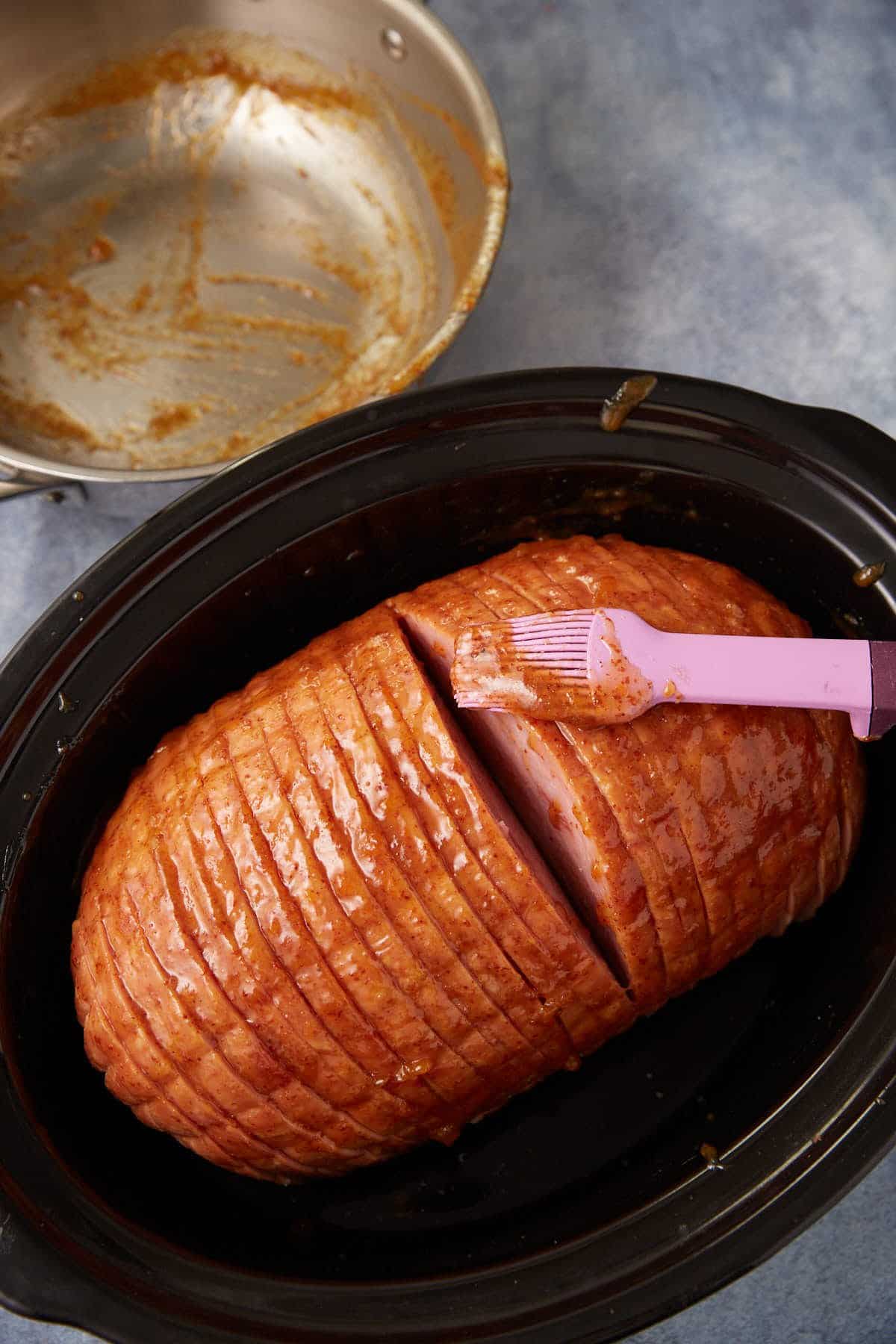 A glazed ham in a black slow cooker is being brushed with sauce using a pink silicone brush. A cut reveals the inside of the ham. A metal pan with leftover glaze sits nearby on a blue surface.