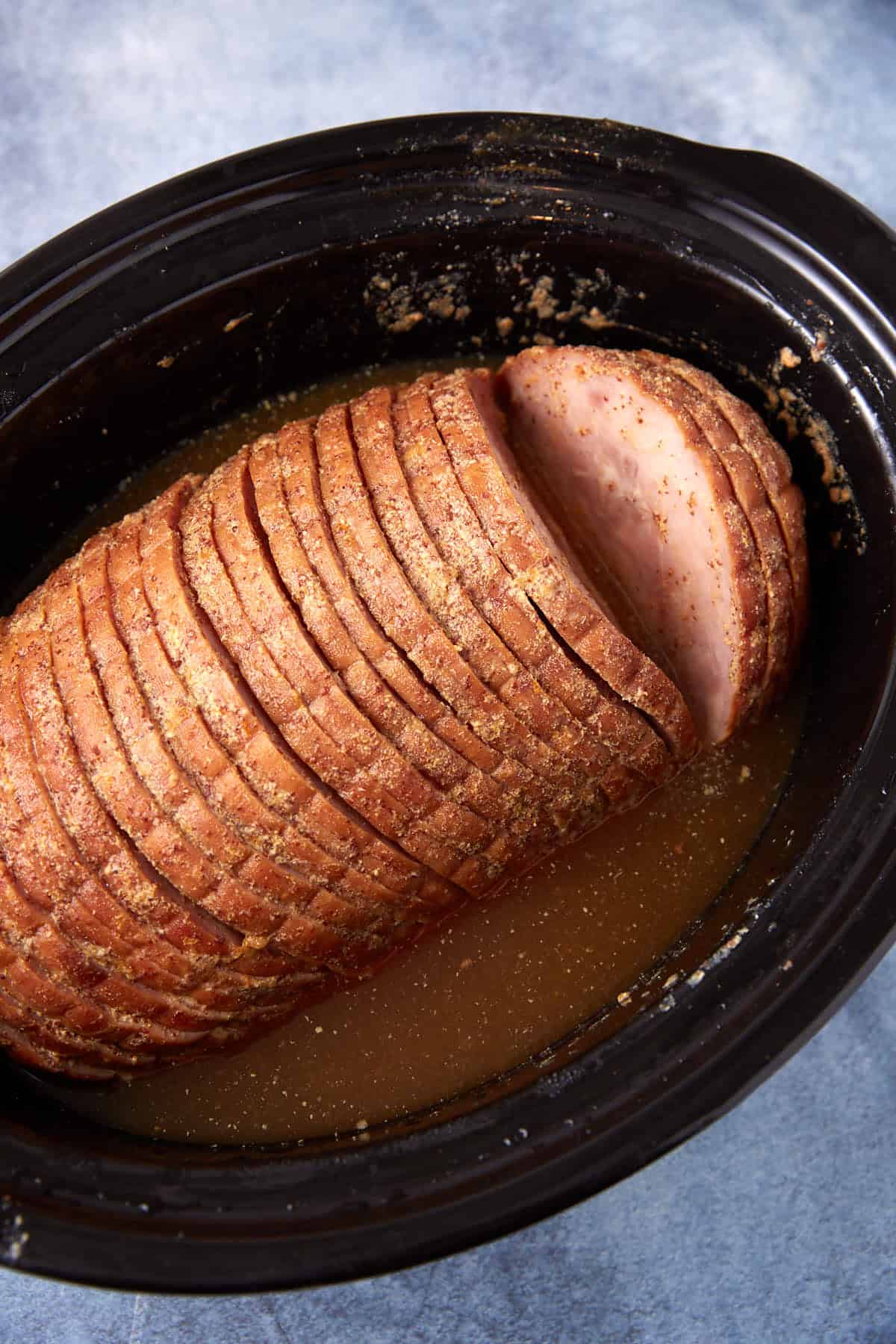 A sliced ham sits in a black slow cooker, surrounded by a light brown liquid and seasonings, on a blue surface.