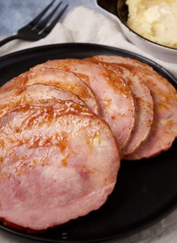 Slices of glazed ham arranged on a black plate, with a fork nearby and a bowl of mashed potatoes in the background.