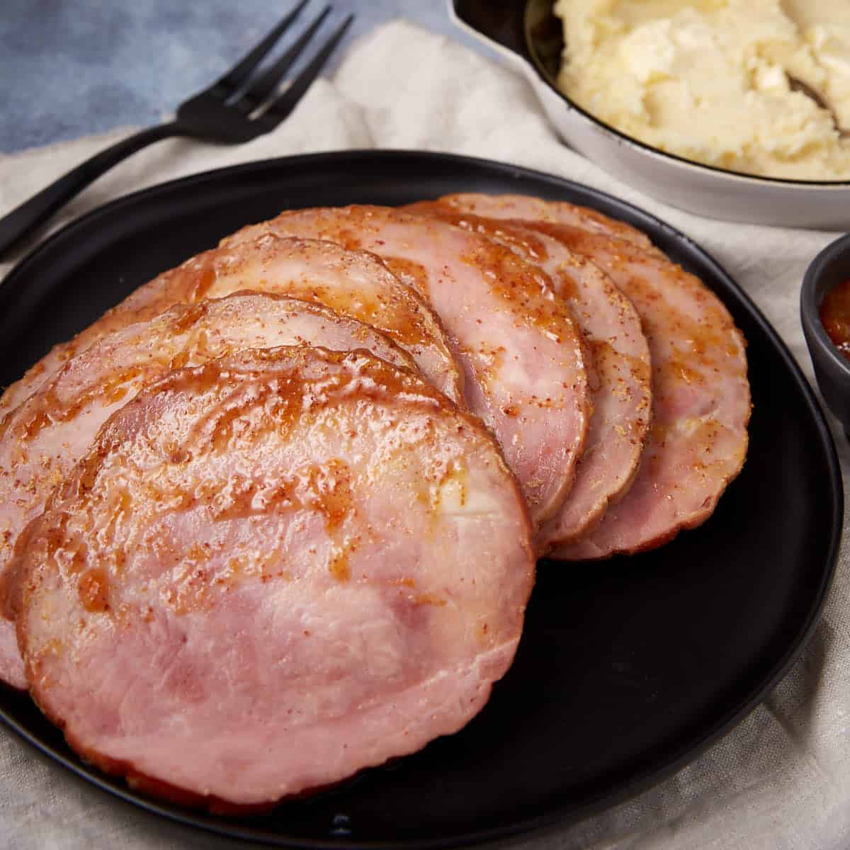 Slices of glazed ham arranged on a black plate, with a fork nearby and a bowl of mashed potatoes in the background.