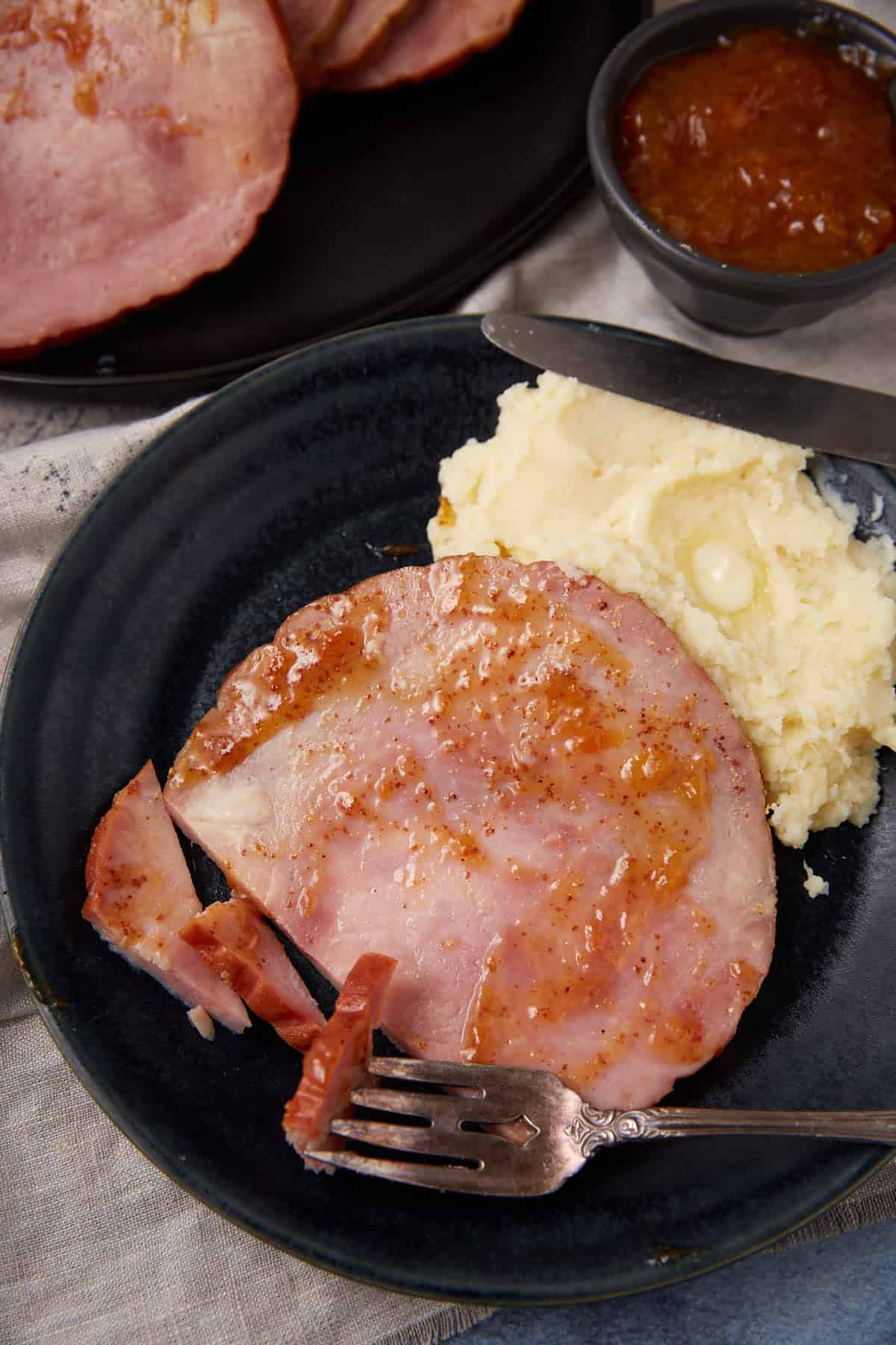 A plate with a slice of glazed ham, mashed potatoes with a pat of butter, and a fork. In the background, there’s a small bowl of sauce and a platter with more ham slices.