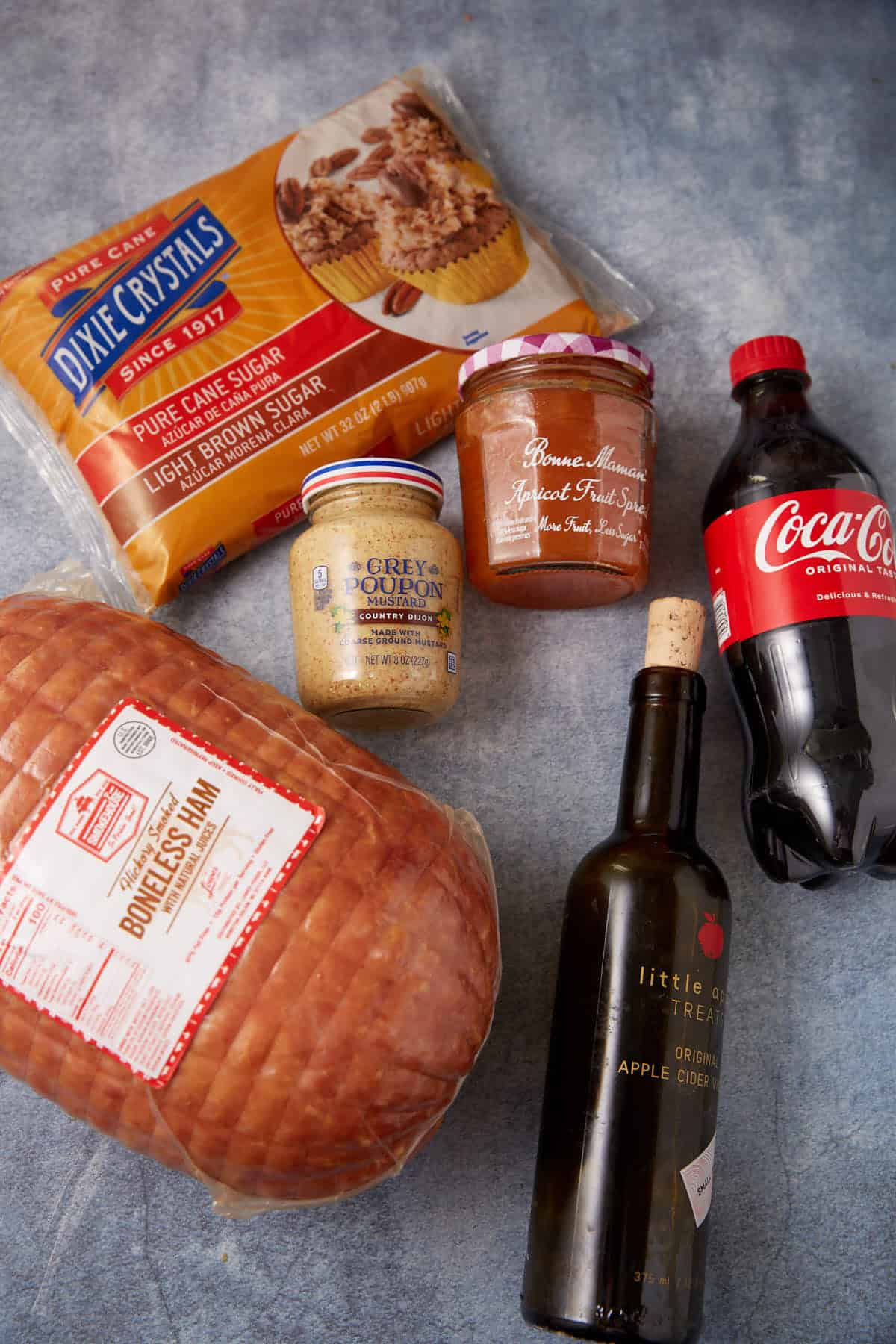 A collection of ingredients on a gray surface: a large ham, a bottle of apple cider, a bottle of Coca-Cola, a bag of light brown sugar, a jar of Dijon mustard, and a jar of apricot preserves.