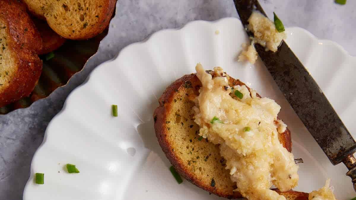 A slice of toasted bread on a white plate is topped with a creamy spread and garnished with chopped green herbs, hinting at the first plates cleared. A knife with some spread rests beside it, while more toasts are visible in a dish nearby.