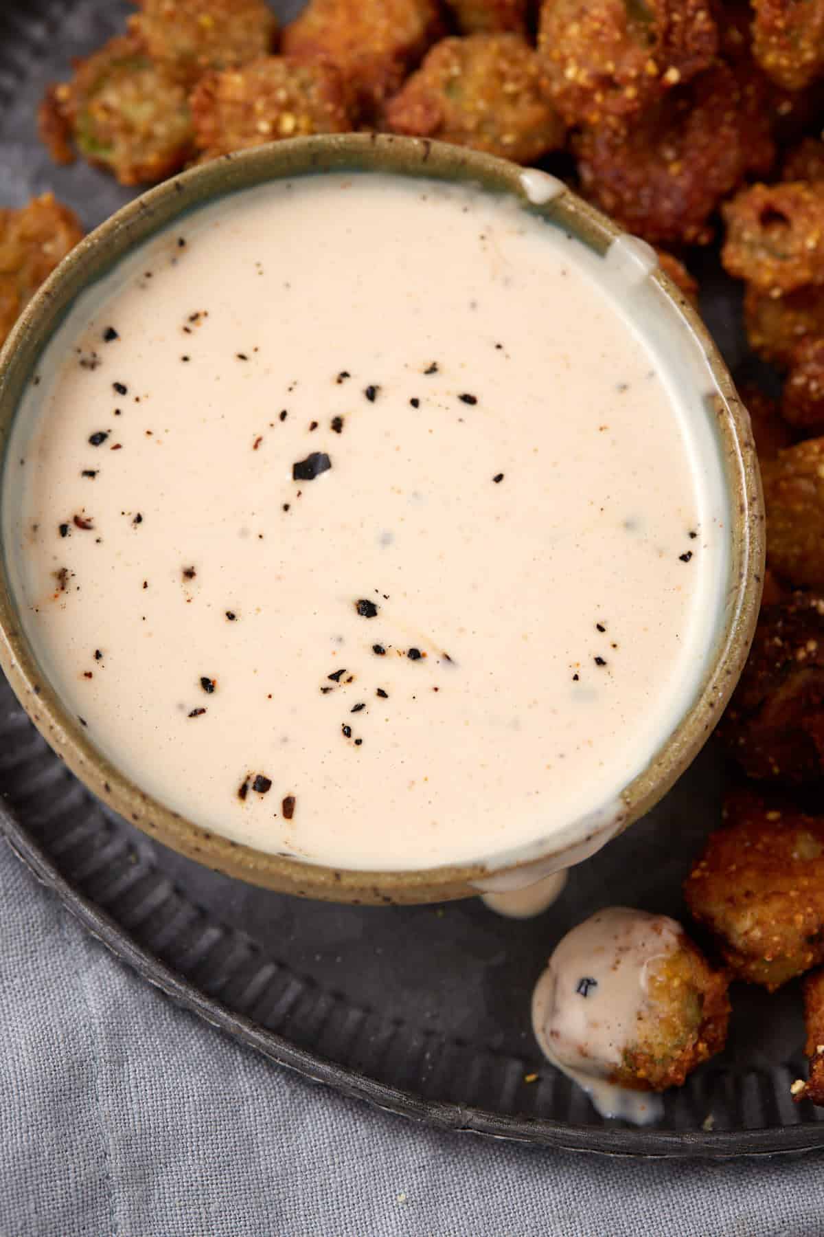 A bowl of creamy dipping sauce with cracked black pepper on top sits on a metal tray next to pieces of fried food, possibly okra. A bit of sauce is dripping down the side of the bowl.