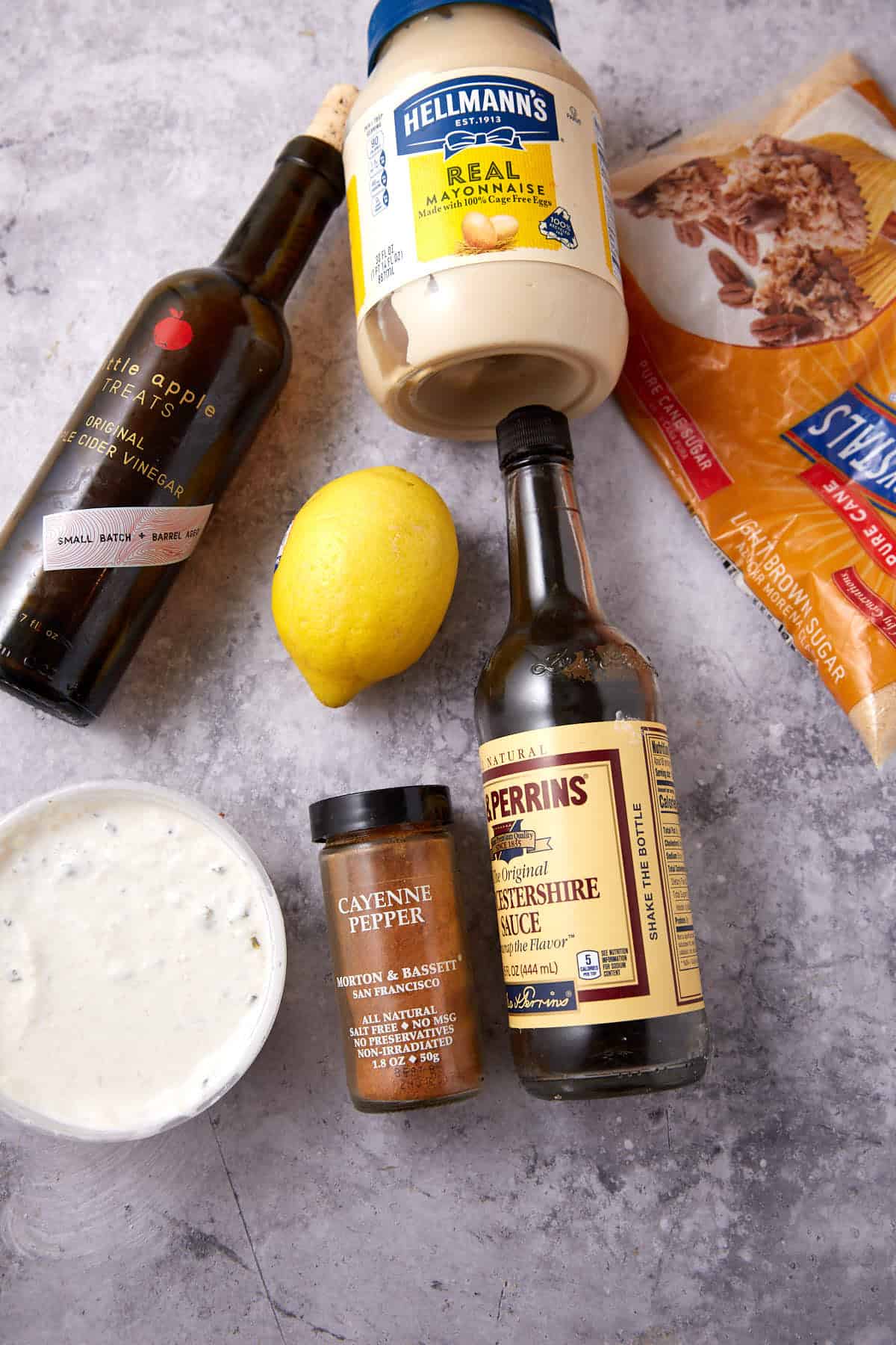 Assorted ingredients on a countertop: a bottle of balsamic vinegar, a jar of mayonnaise, a lemon, a bottle of Worcestershire sauce, a container of cayenne pepper, a bowl of dressing, and a bag of brown sugar.