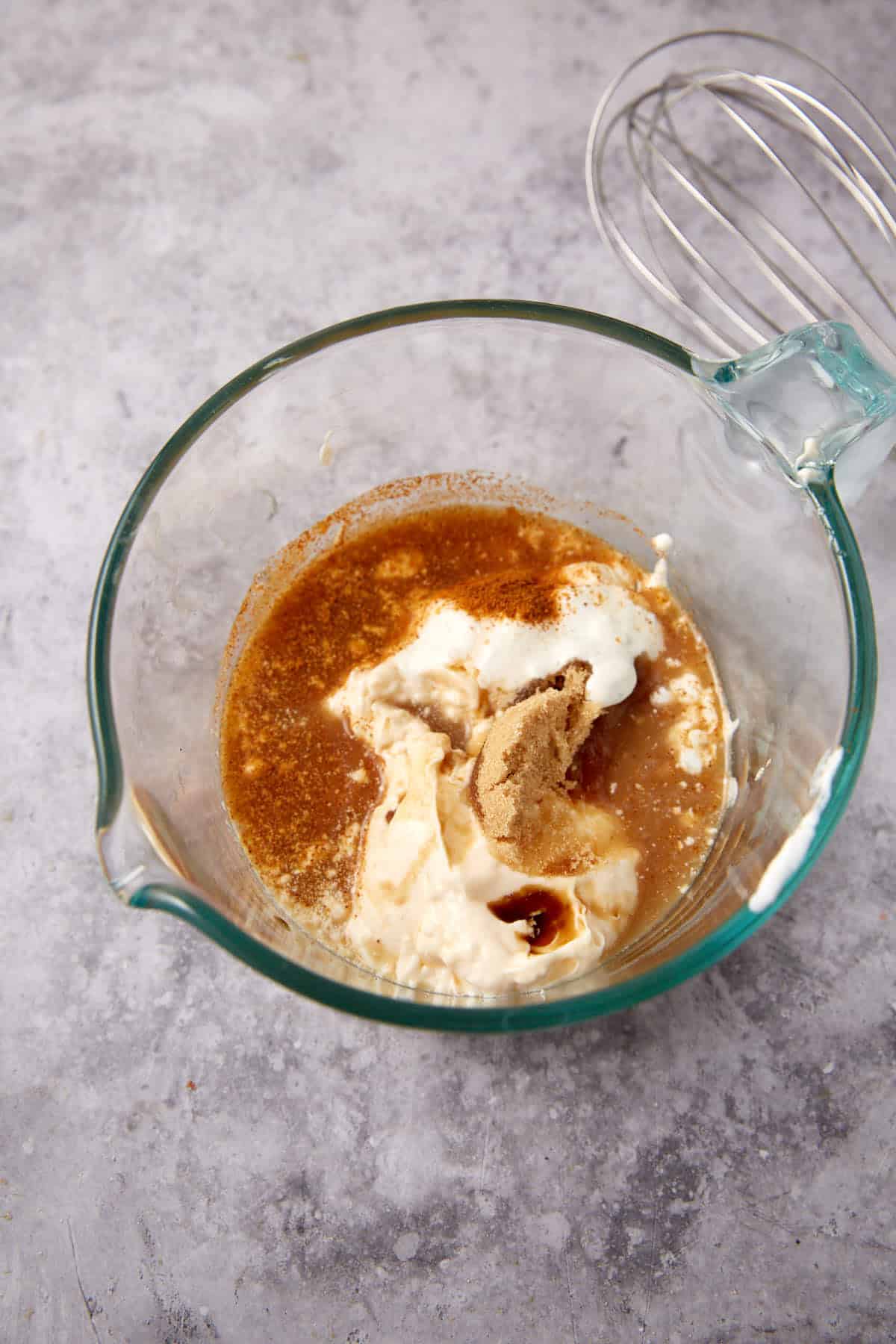 A glass mixing bowl with brown sugar, mayonnaise, sour cream, spices, and liquid ingredients being prepared on a gray countertop. A metal whisk is resting nearby.