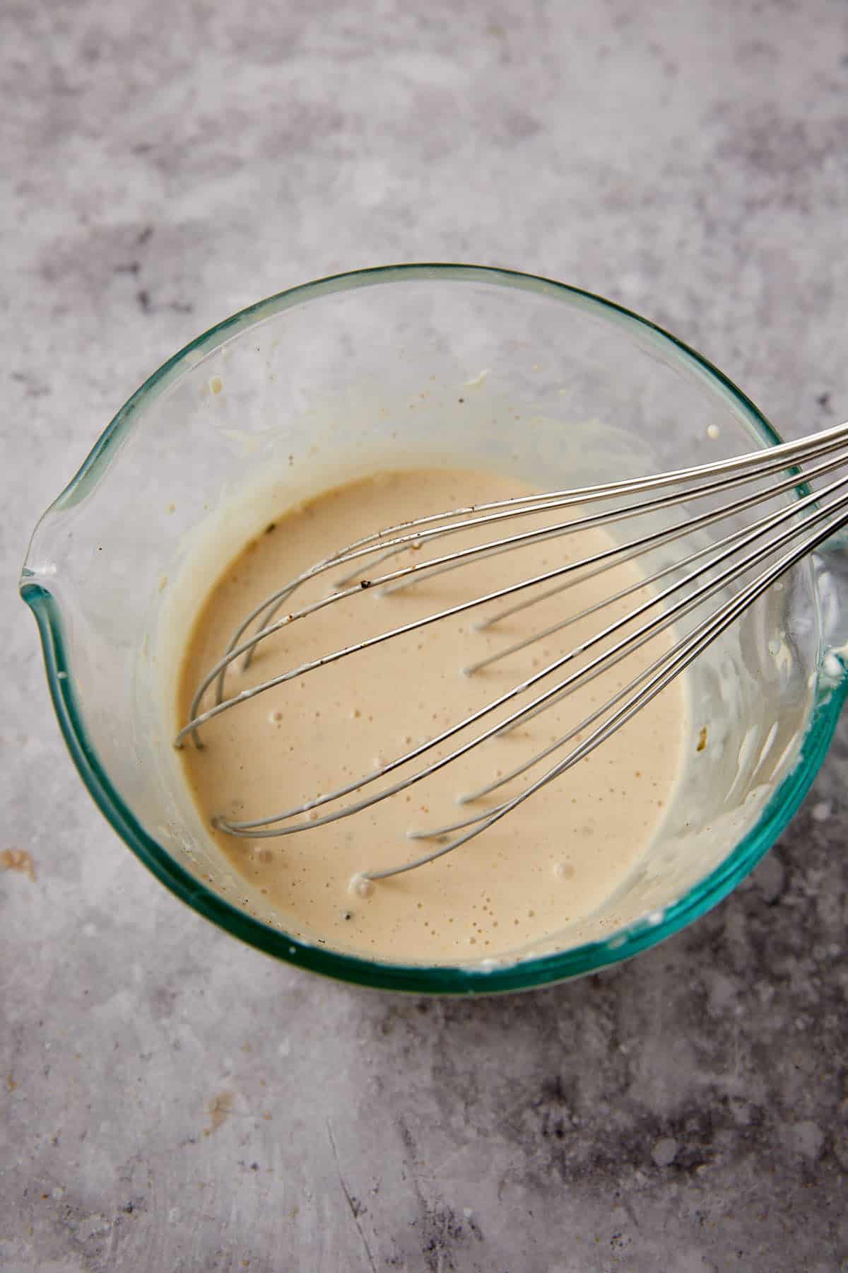 A large metal whisk rests in a glass mixing bowl containing a light tan batter or mixture, set on a textured gray surface.