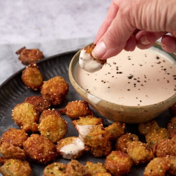 A hand dips a piece of fried pickle into a bowl of creamy sauce sprinkled with black pepper. More fried pickles are scattered on a dark plate beside the bowl.