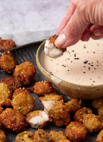 A hand dips a piece of fried pickle into a bowl of creamy sauce sprinkled with black pepper. More fried pickles are scattered on a dark plate beside the bowl.