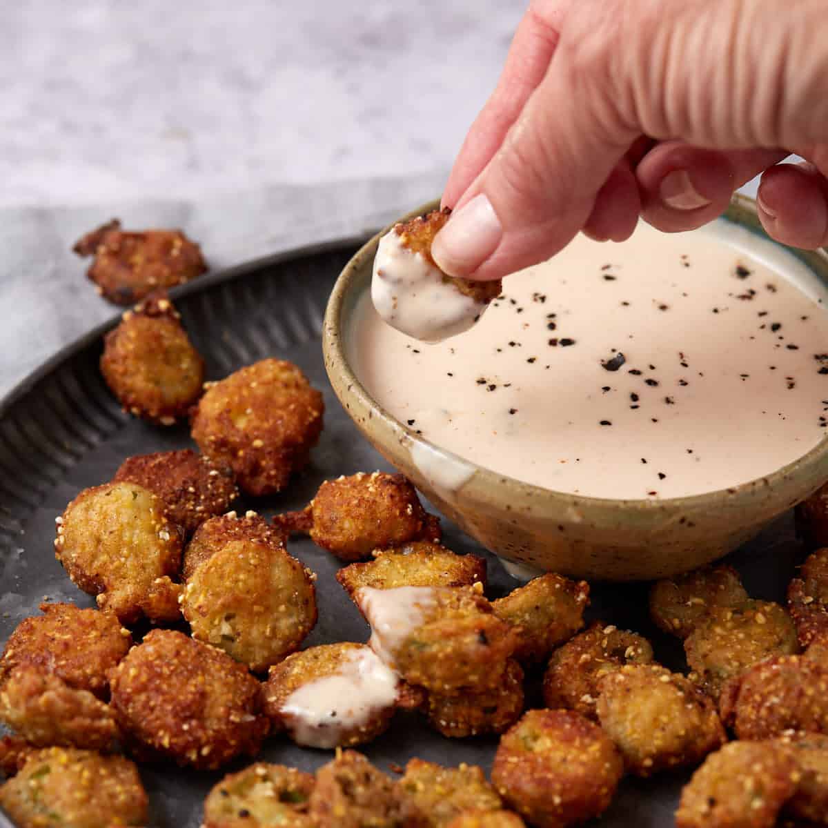 A hand dips a piece of fried pickle into a bowl of creamy sauce sprinkled with black pepper. More fried pickles are scattered on a dark plate beside the bowl.