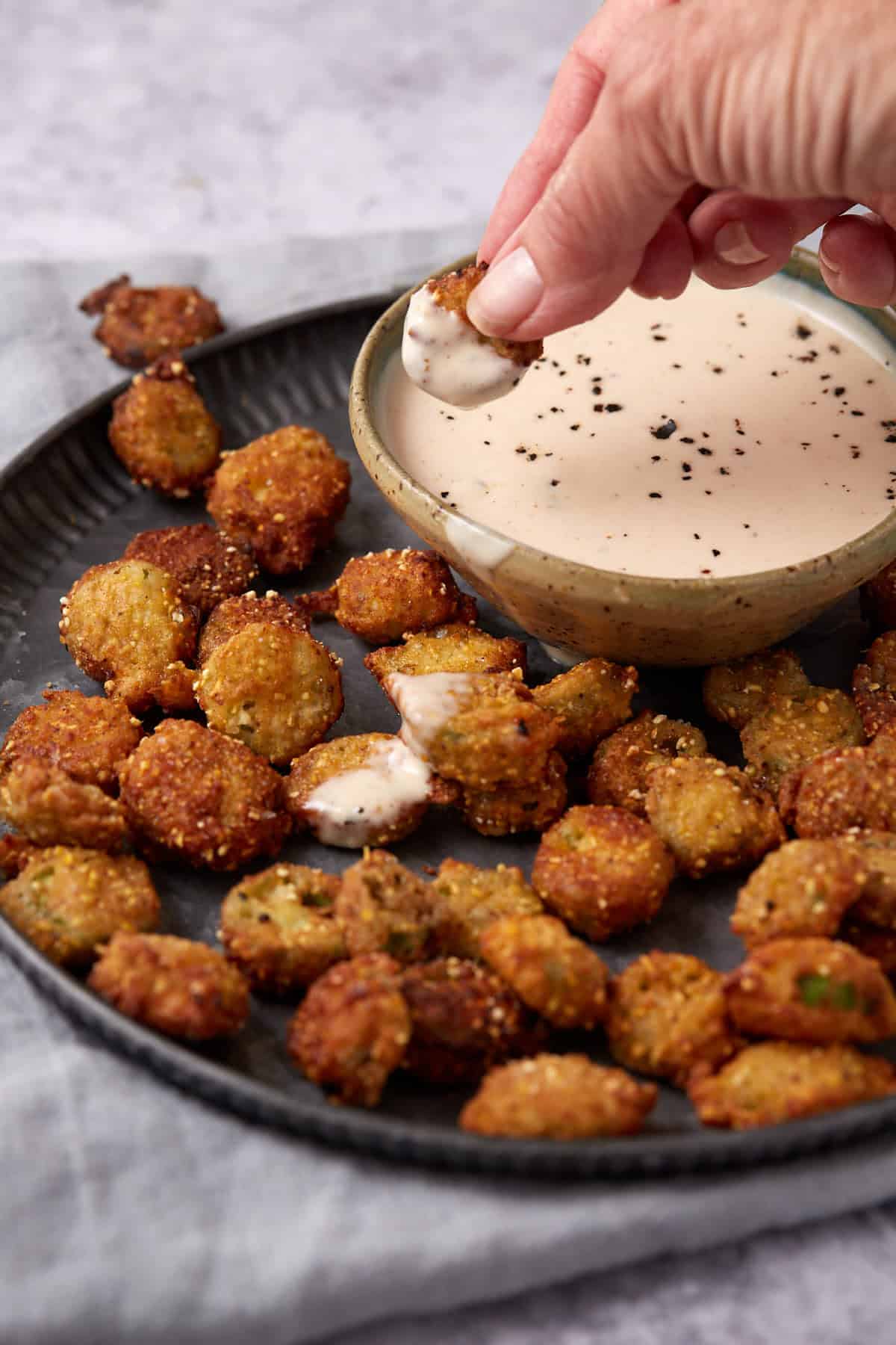 A hand dips a piece of fried okra into a bowl of creamy dipping sauce on a dark plate filled with more pieces of fried okra.