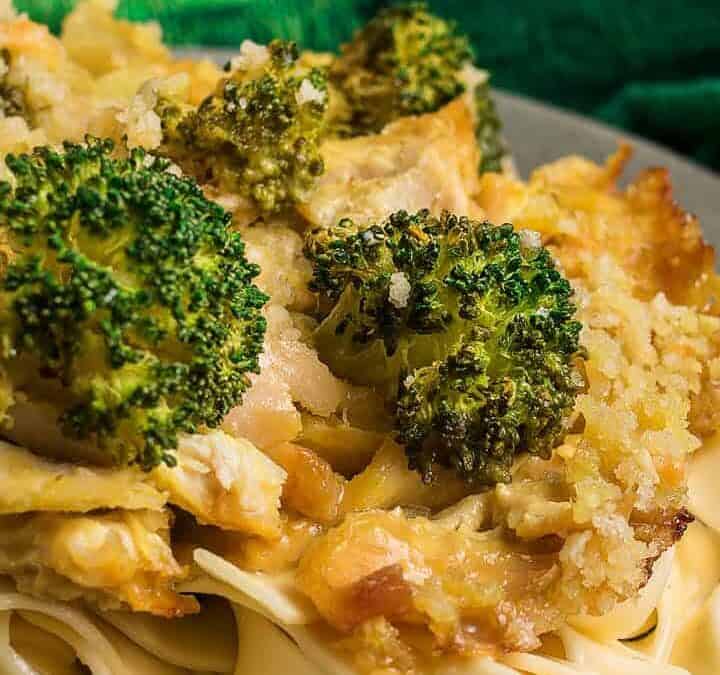 A plate of wide, flat pasta topped with a cheesy Chicken Divan broccoli and breadcrumb casserole, with a baking dish of the casserole in the background.