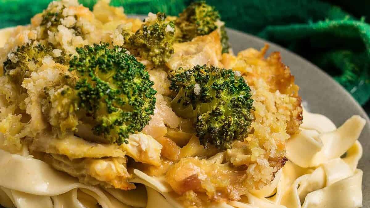 A plate of wide, flat pasta topped with a cheesy Chicken Divan broccoli and breadcrumb casserole, with a baking dish of the casserole in the background.
