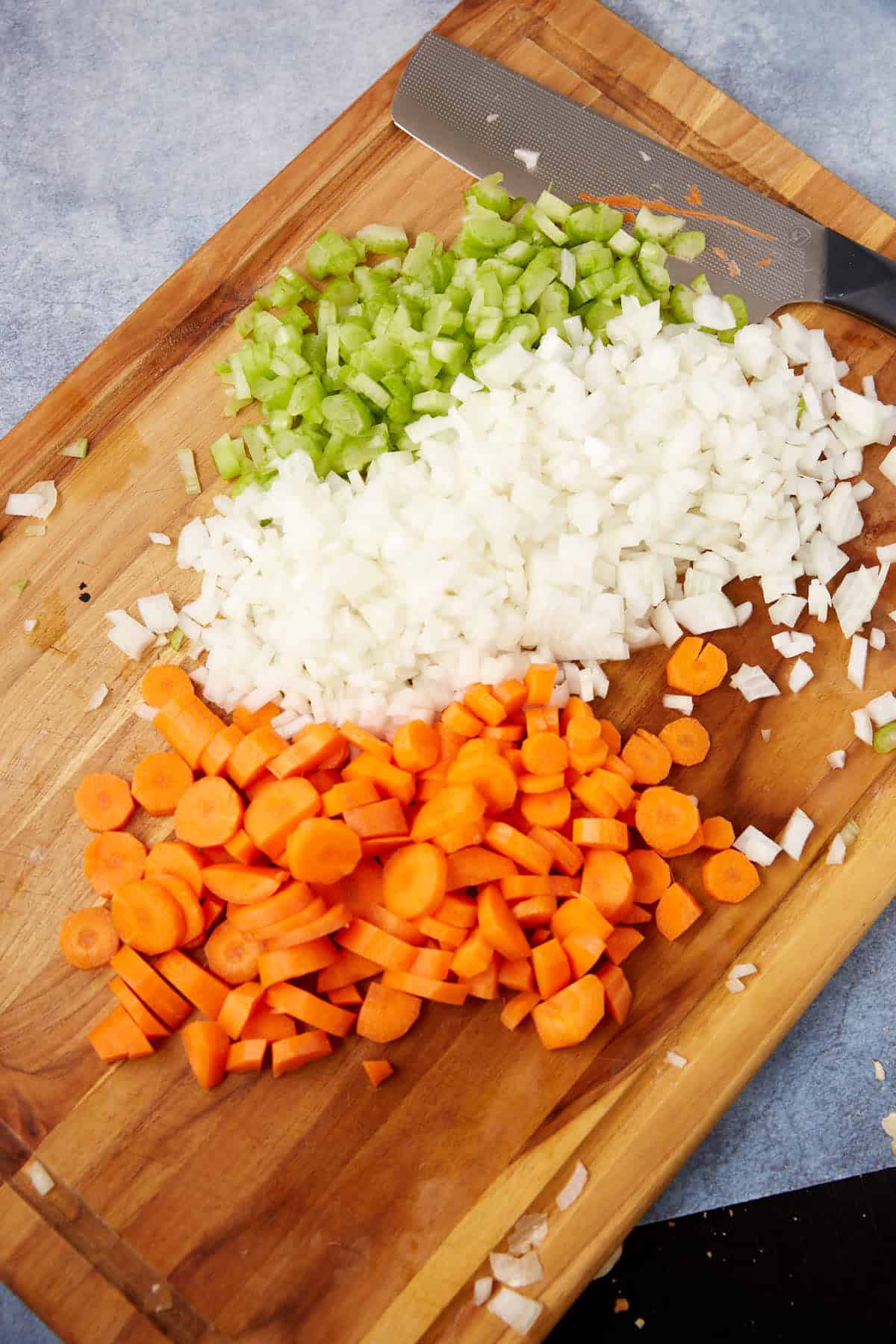 A wooden cutting board with chopped celery, diced onion, and sliced carrots arranged in rows, next to a knife.