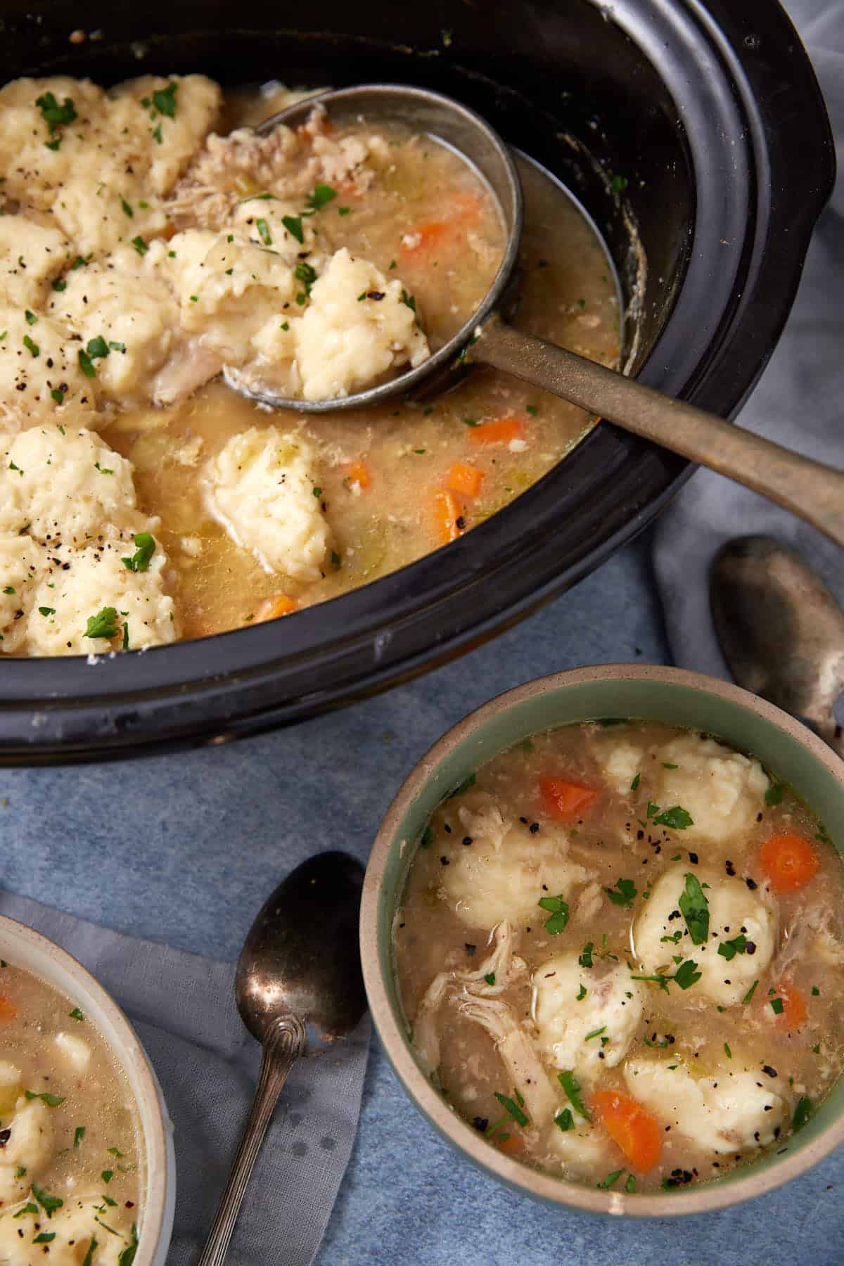 A bowl and slow cooker filled with chicken and dumplings soup, featuring chunks of chicken, vegetables, fluffy dumplings, and garnished with chopped parsley, with spoons and a napkin beside them.