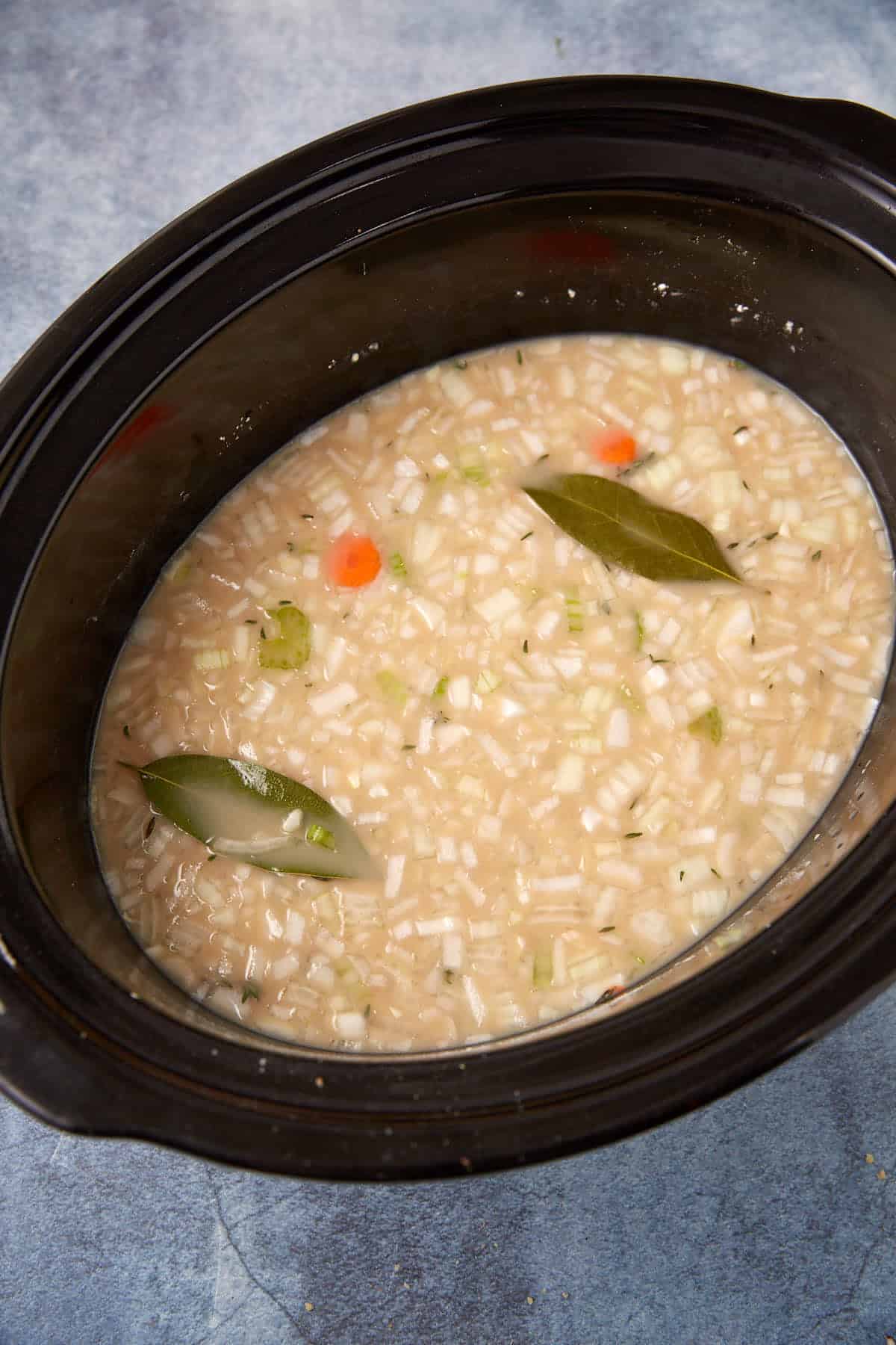 A slow cooker filled with uncooked soup ingredients, including chopped onions, celery, carrots, rice, bay leaves, and broth, on a textured blue surface.