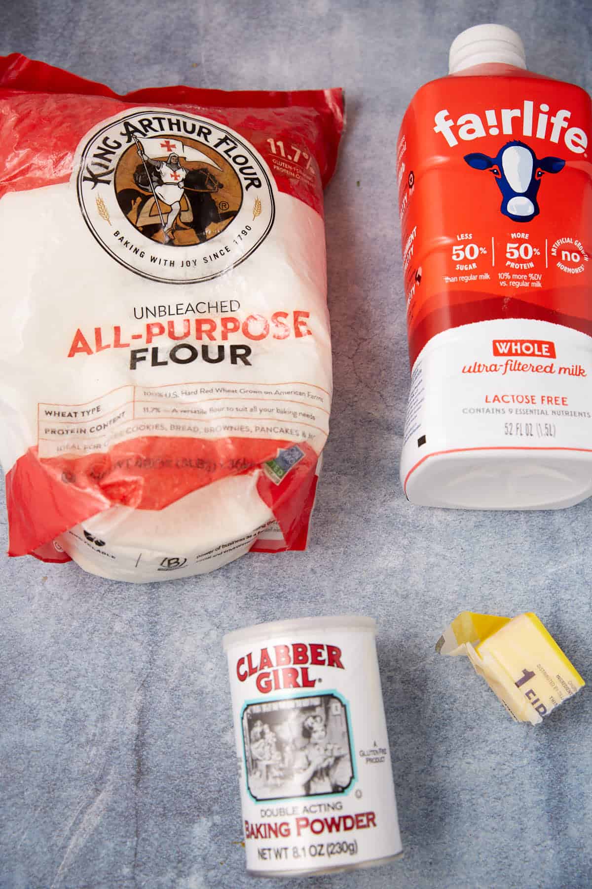 A bag of King Arthur all-purpose flour, a carton of Fairlife whole milk, a can of Clabber Girl baking powder, and a small portion of butter on a light blue textured surface.