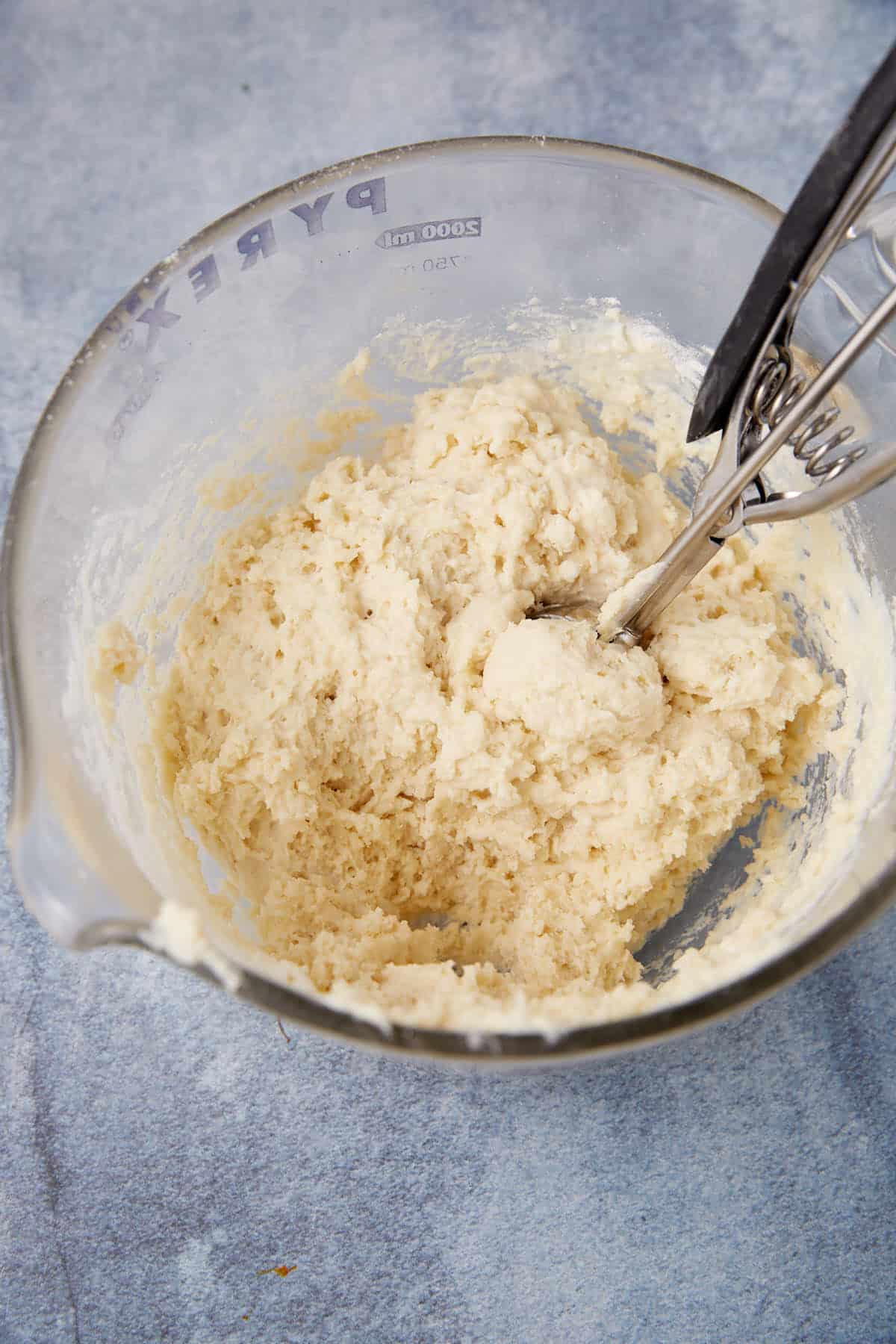 A glass mixing bowl filled with pale, sticky biscuit dough and a metal cookie scoop resting inside, all on a light blue textured surface.