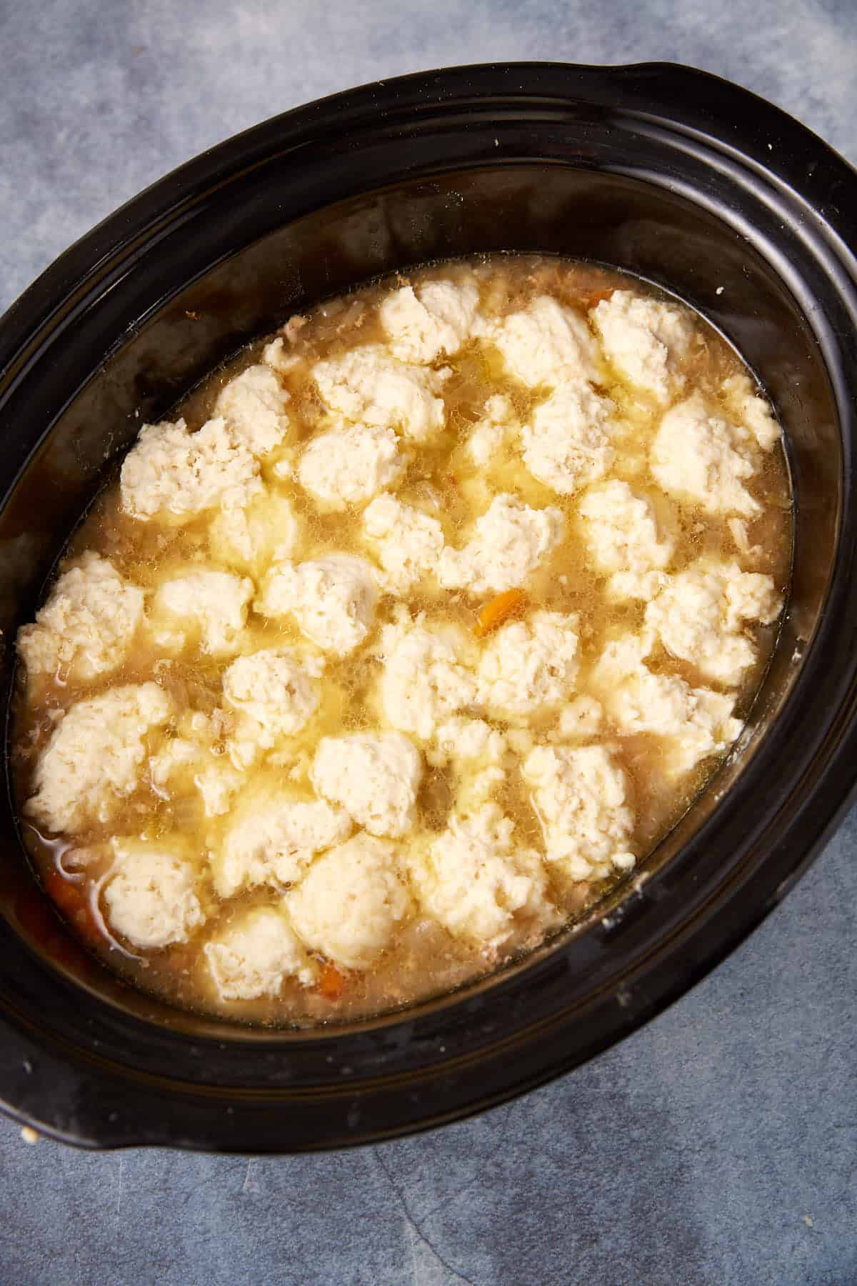 A slow cooker filled with chicken and dumplings; round dumplings are floating in a thick, golden broth with visible bits of chicken and vegetables.