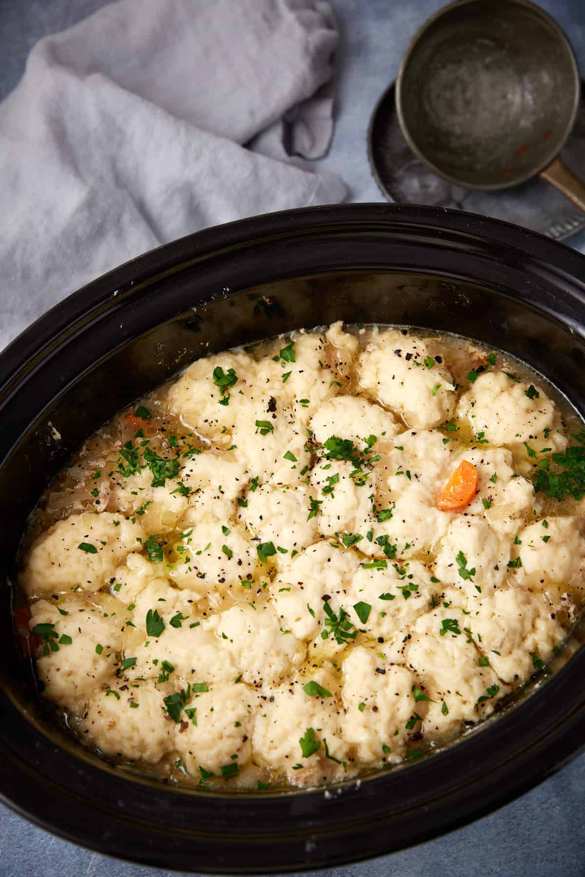 A slow cooker filled with chicken and dumplings stew, topped with fluffy dumplings and garnished with chopped parsley. A gray cloth and a ladle are in the background.