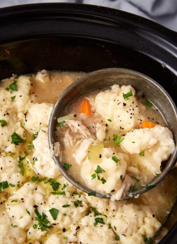 A ladle scoops up chicken and dumplings with vegetables from a slow cooker. The dish is creamy, topped with parsley, and contains chunks of carrots, celery, shredded chicken, and fluffy dumplings.