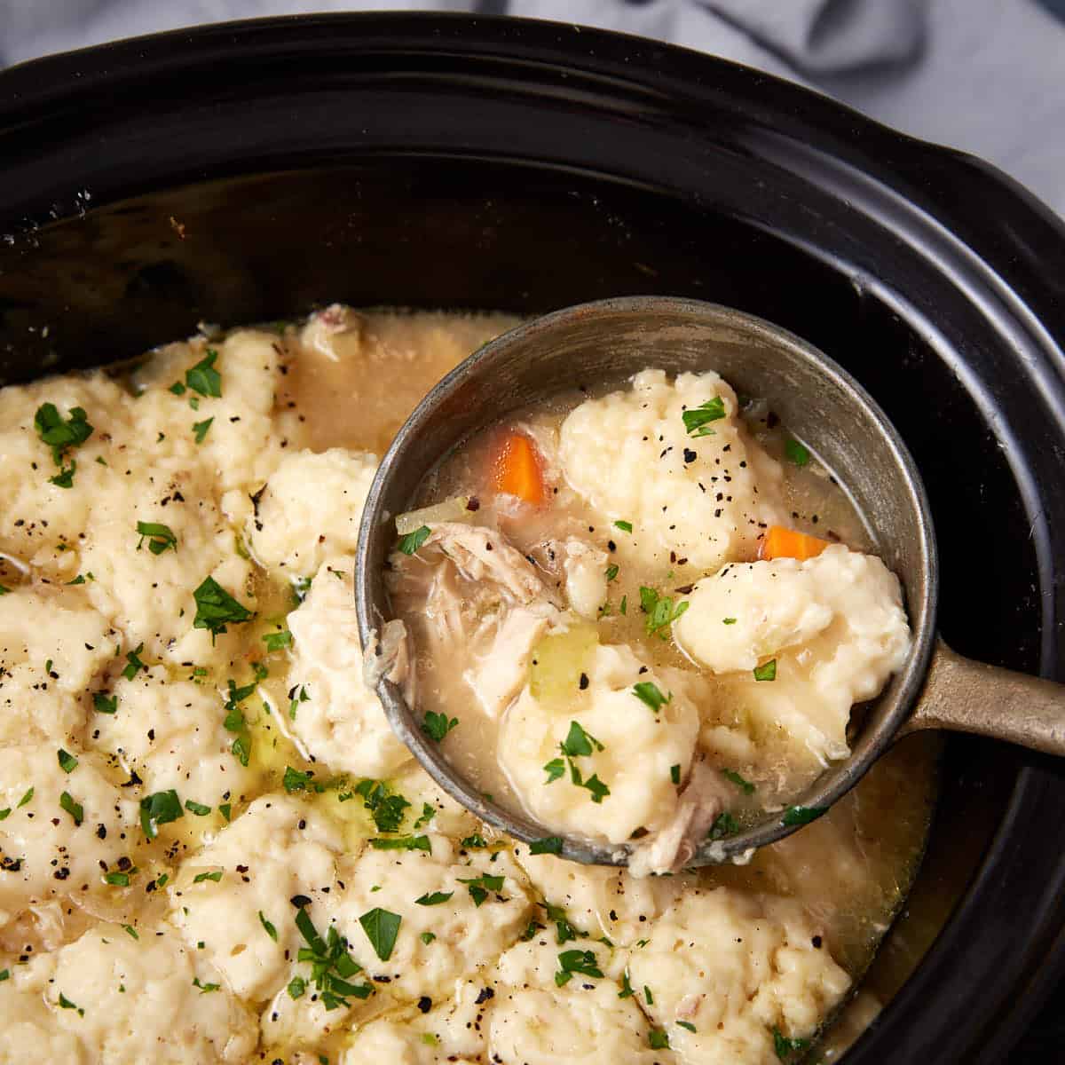 A ladle scoops up chicken and dumplings with vegetables from a slow cooker. The dish is creamy, topped with parsley, and contains chunks of carrots, celery, shredded chicken, and fluffy dumplings.