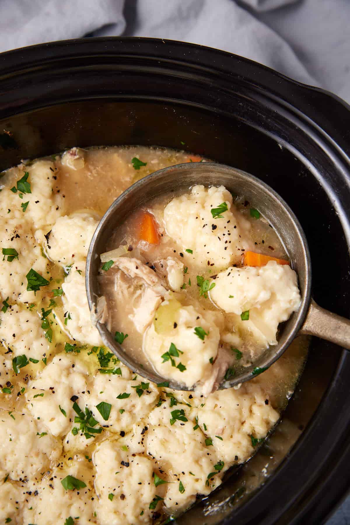 A ladle scoops chicken and dumplings from a slow cooker, showing fluffy dumplings, shredded chicken, carrots, celery, broth, and chopped parsley garnish.
