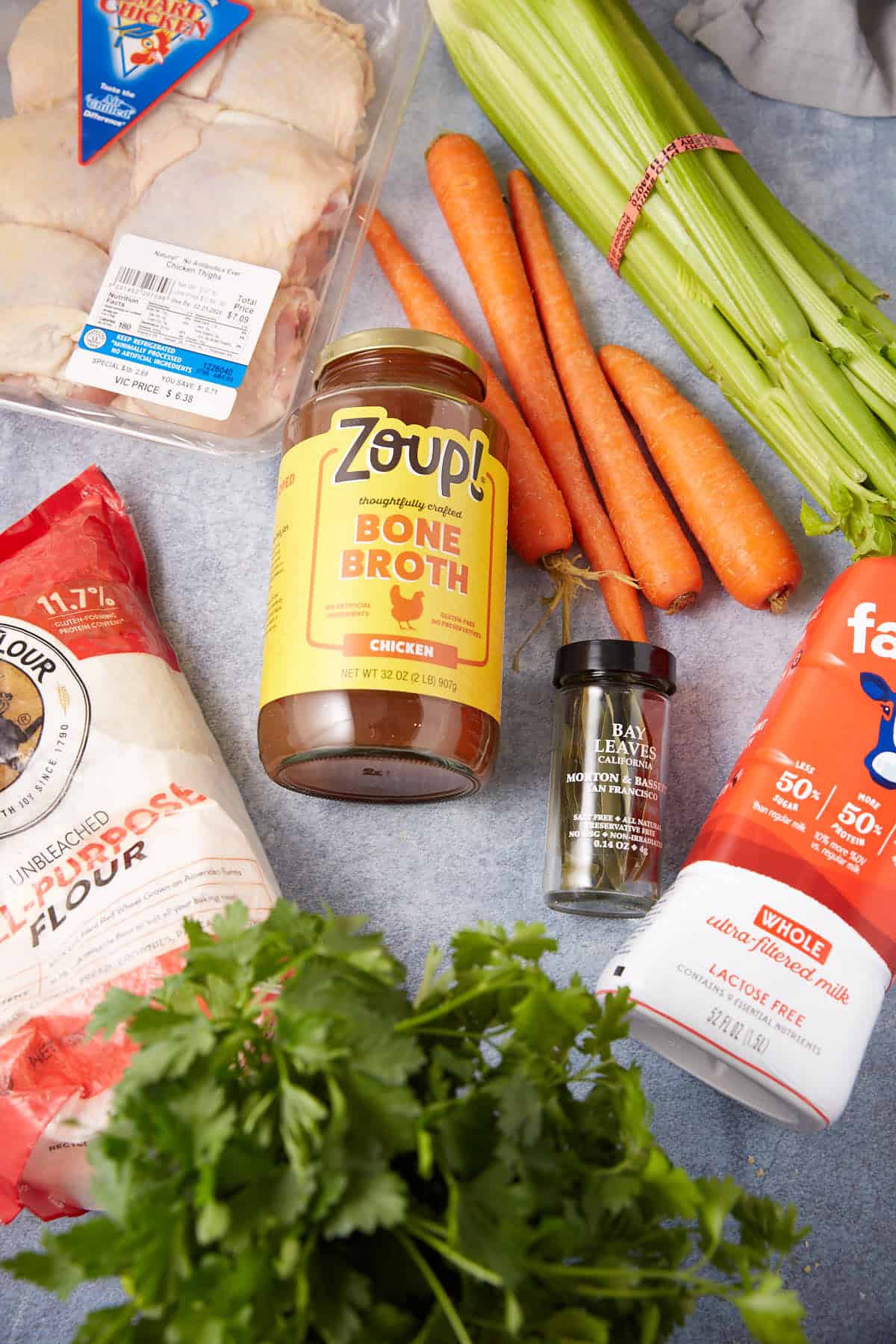 A top-down view of ingredients on a countertop: raw chicken, carrots, celery, a jar of bone broth, a bottle of dried bay leaves, a bunch of parsley, flour, and a container of lactose-free milk.