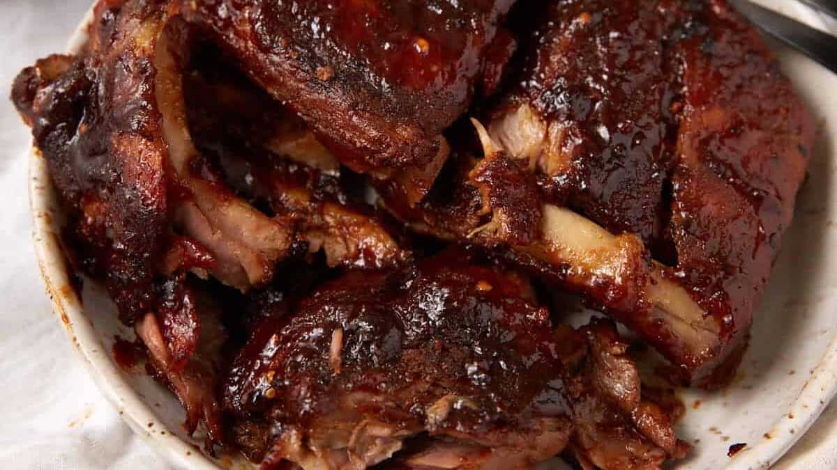 A plate of saucy, glazed slow cooker ribs sits on a cloth with a bowl of macaroni and cheese and a dish of barbecue sauce nearby. A pink basting brush and fork are also visible.