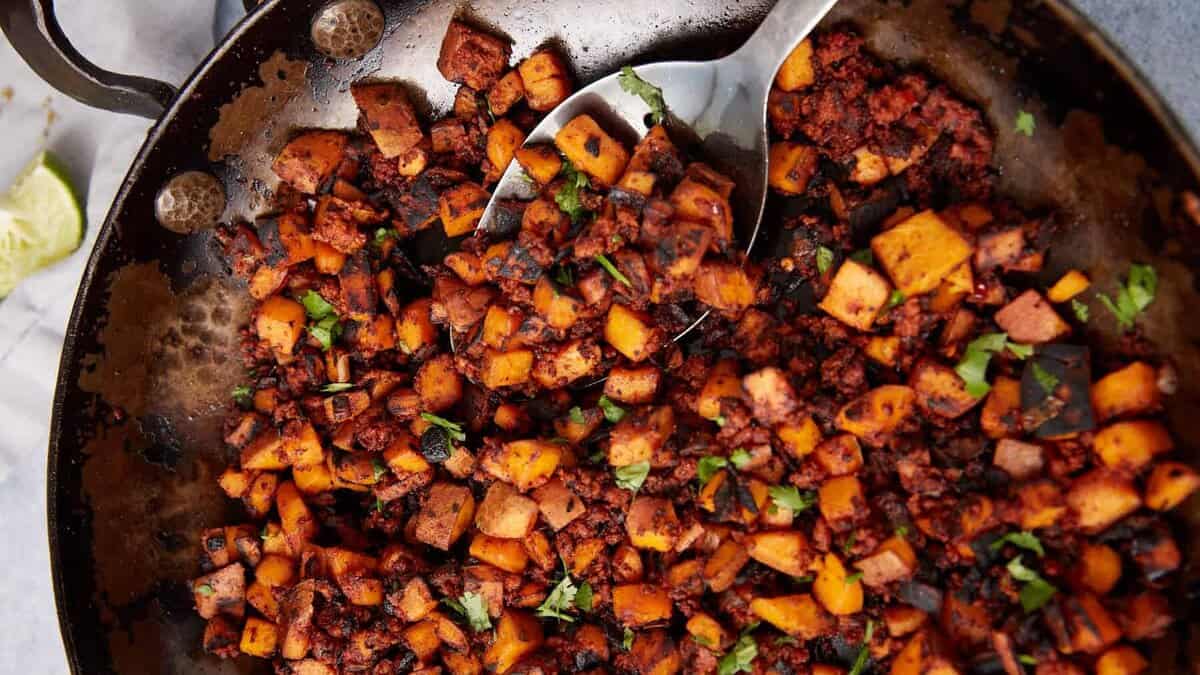 A skillet filled with cooked diced vegetables and crumbled chorizo, garnished with fresh herbs. This vibrant Sweet Potato Hash features orange, brown, and green tones, with a metal spoon resting in the pan, ready to serve.