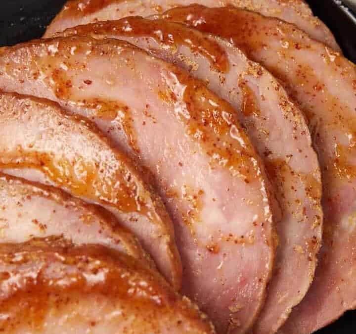 Sliced glazed ham arranged on a black plate with a side of mashed potatoes in a bowl and a small dish of sauce, all on a light-colored cloth.