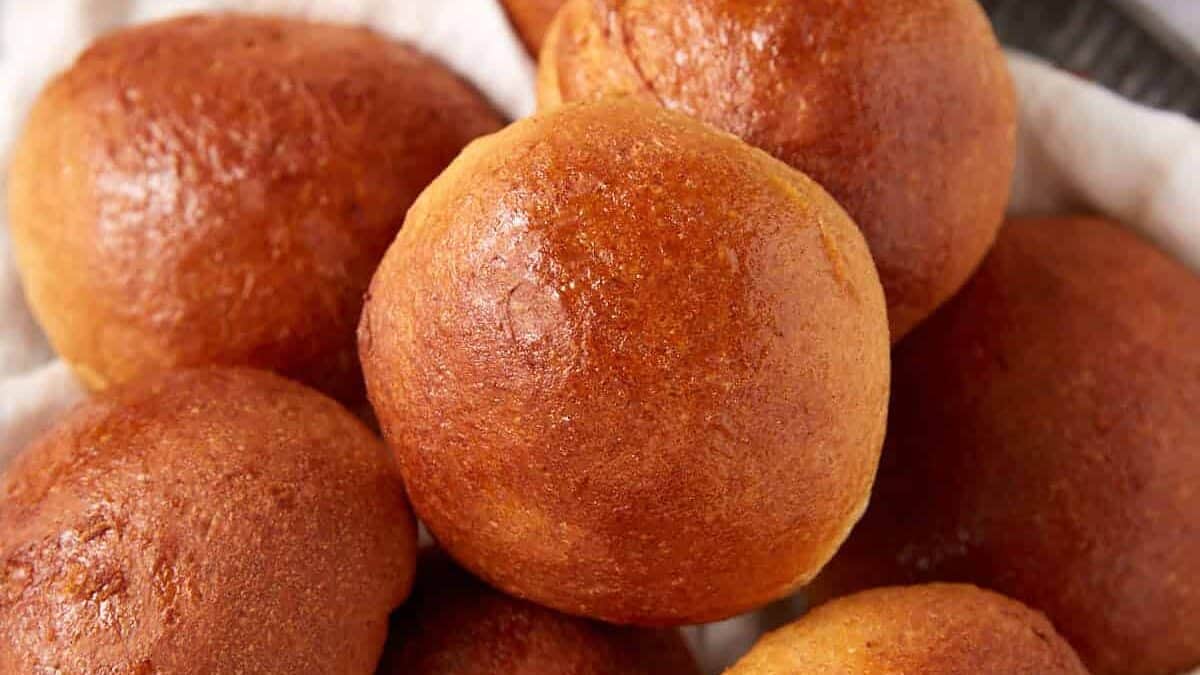 A plate of golden brown whole wheat rolls sits on a white cloth, with a dish of honey and a honey dipper in the background.