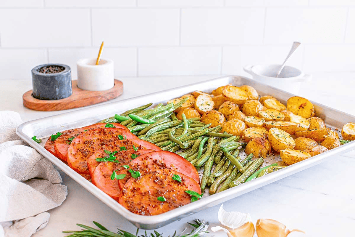 A sheet pan with slices of ham, green beans, and roasted baby potatoes garnished with herbs shows how sheet pan dinners keep cleanup simple, set on a white countertop with salt and pepper containers in the background.
