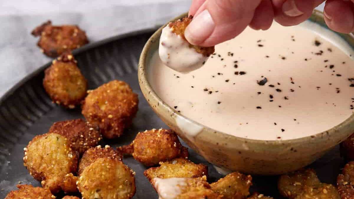 A hand dips a piece of fried okra into a bowl of creamy dipping sauce on a dark plate 