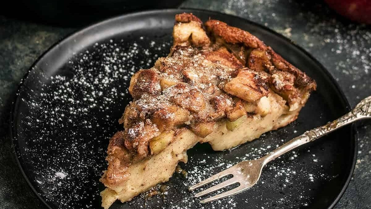 A German apple pancake dessert dusted with powdered sugar sits in a skillet on a dark cloth