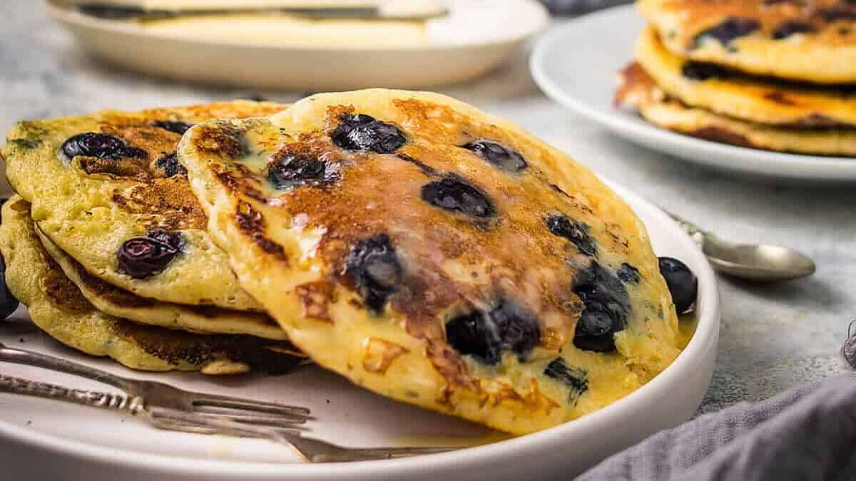 A plate of fluffy blueberry pancakes sits on a table with a fork and knife, accompanied by a cup, a dish of butter with a knife, and a bowl of fresh blueberries in the background. A folded napkin lies neatly in the foreground.