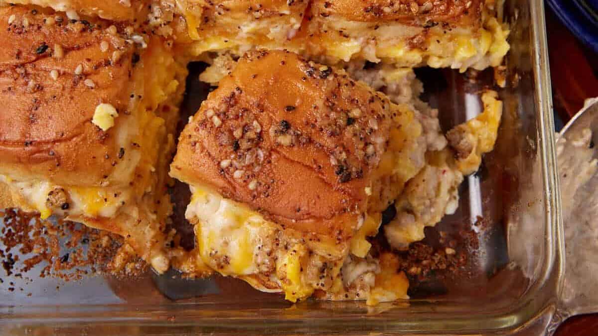 Golden brown breakfast sliders filled with scrambled eggs, cheese, and sausage, topped with a peppery seasoning, arranged in a glass baking dish on a wooden surface—perfect alongside a classic German apple pancake for a hearty breakfast spread.