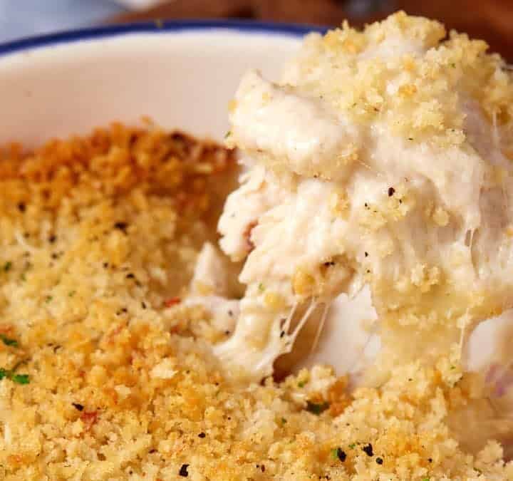 A large blue baking dish filled with a creamy chicken cordon bleu casserole, topped with golden, crispy breadcrumbs. A serving spoon lifts out a portion, revealing melted cheese and a rich, creamy filling.