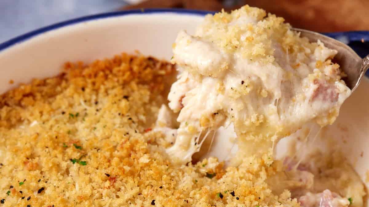 A large blue baking dish filled with a creamy chicken cordon bleu casserole, topped with golden, crispy breadcrumbs. A serving spoon lifts out a portion, revealing melted cheese and a rich, creamy filling.