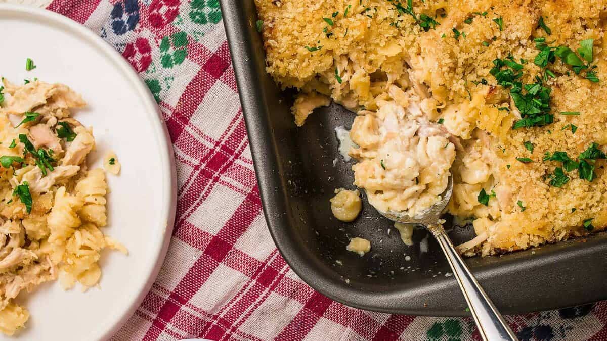 A baking dish of cheesy chicken noodle casserole with a breadcrumb topping, served on a plate above. A spoon rests in the dish, and a small bowl of shredded cheese sits nearby on a red and white checkered cloth.