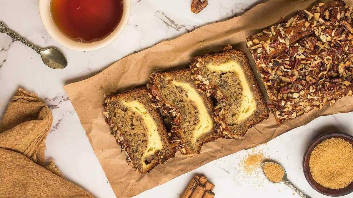 A sliced loaf of Cream Cheese Filled Banana Bread sits on brown paper. Nearby are a bowl of brown sugar, a spoon, cinnamon sticks, a cup of tea, and a beige napkin arranged on a marble surface.