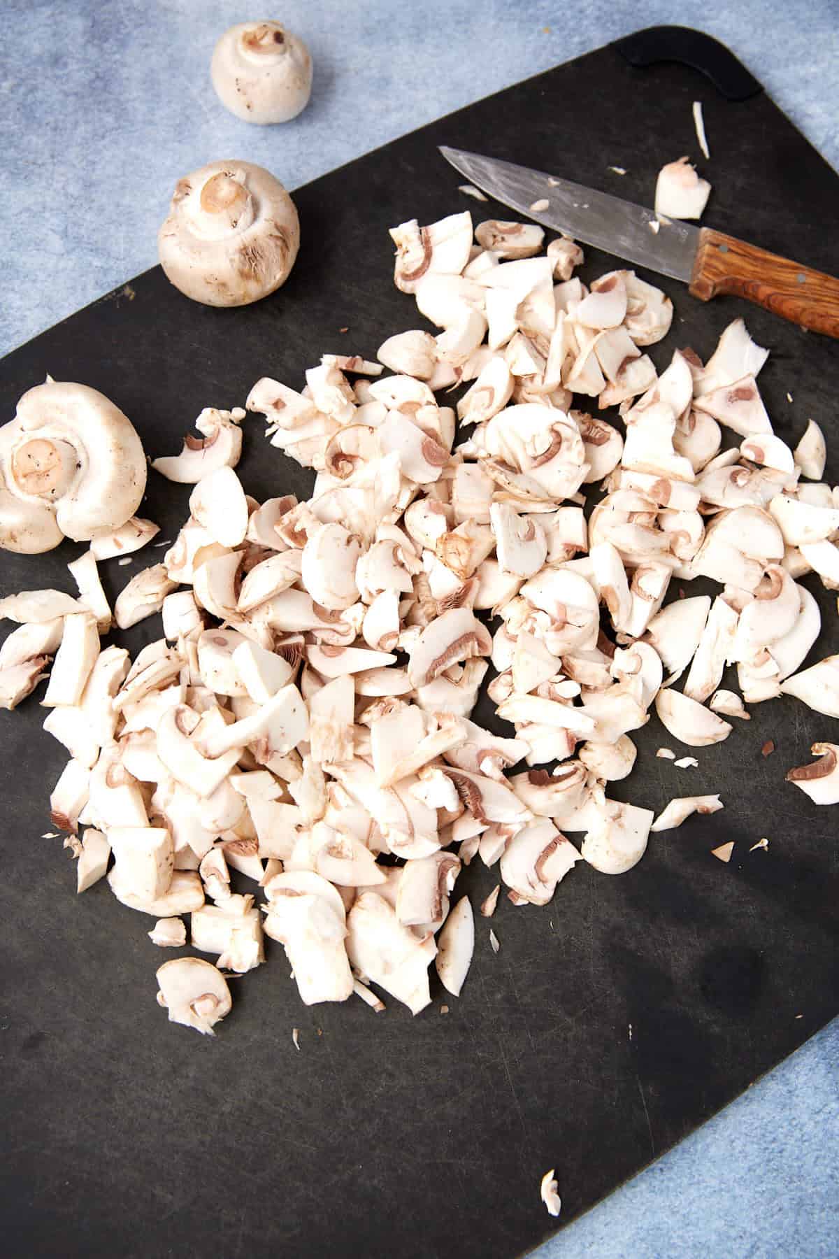 A black cutting board with chopped white mushrooms, a few whole mushrooms, and a knife with a brown handle on a light blue surface.