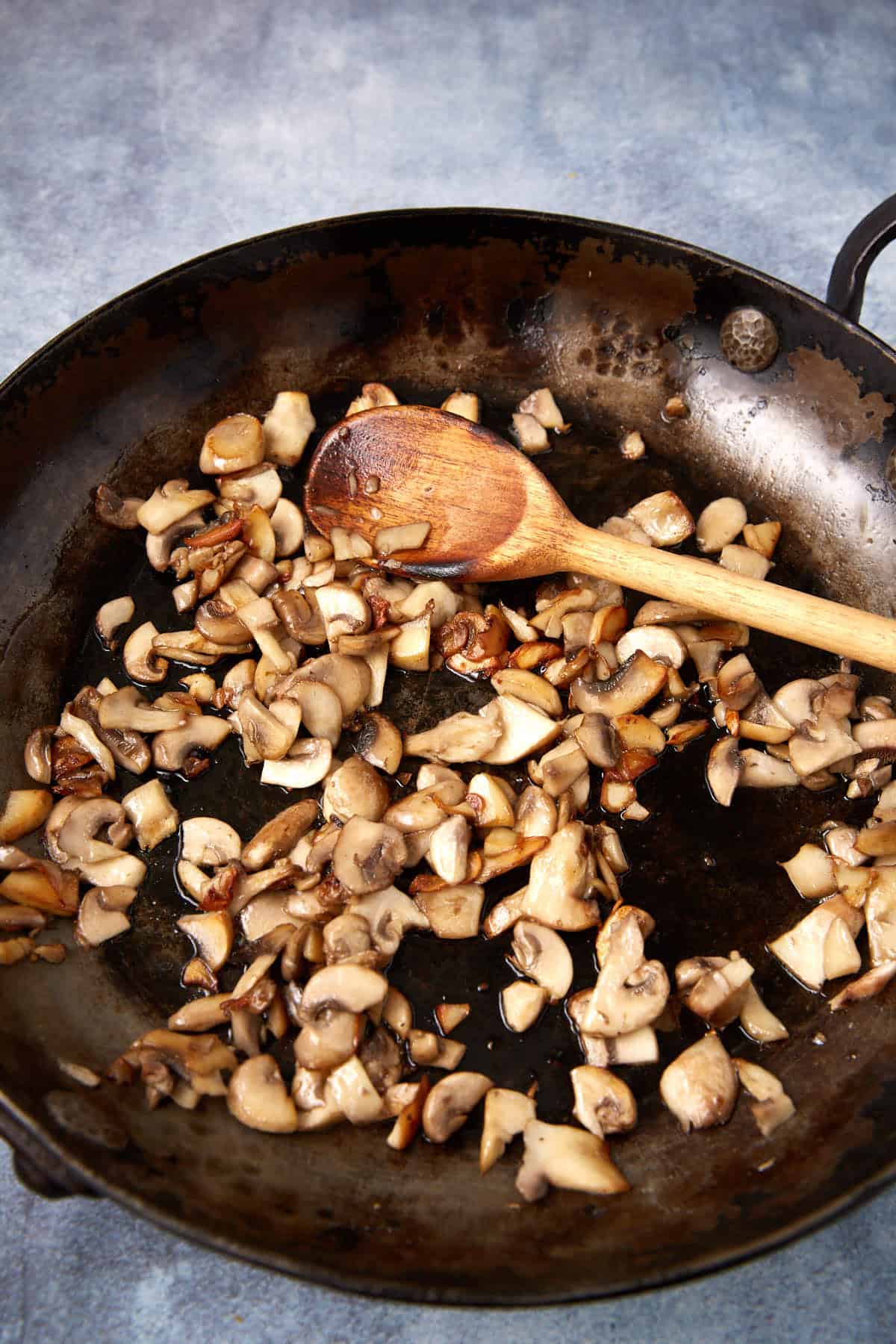 Chopped mushrooms being sautéed in a large, dark skillet with a wooden spoon on a gray surface. The mushrooms are lightly browned and spread across the pan.