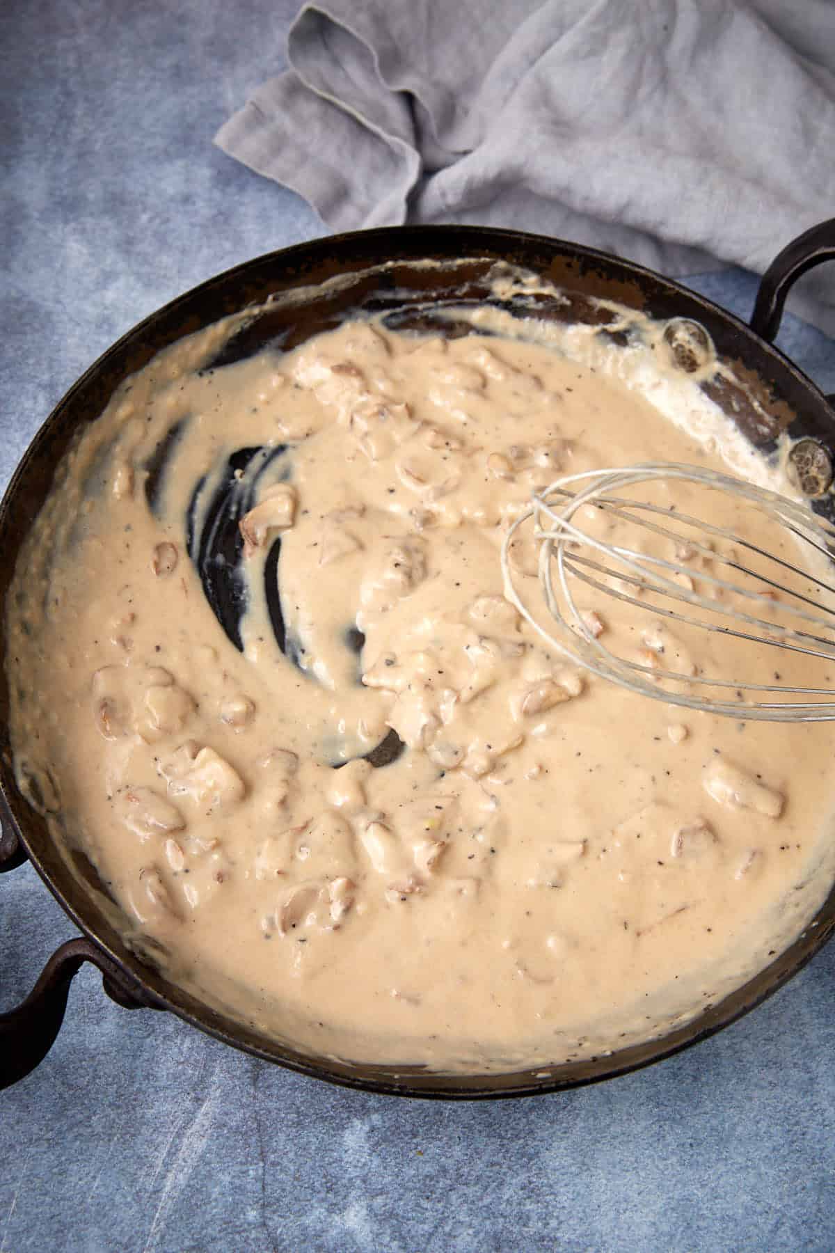 A skillet filled with creamy gravy containing small pieces of sausage, with a metal whisk resting on the side and a gray cloth in the background.